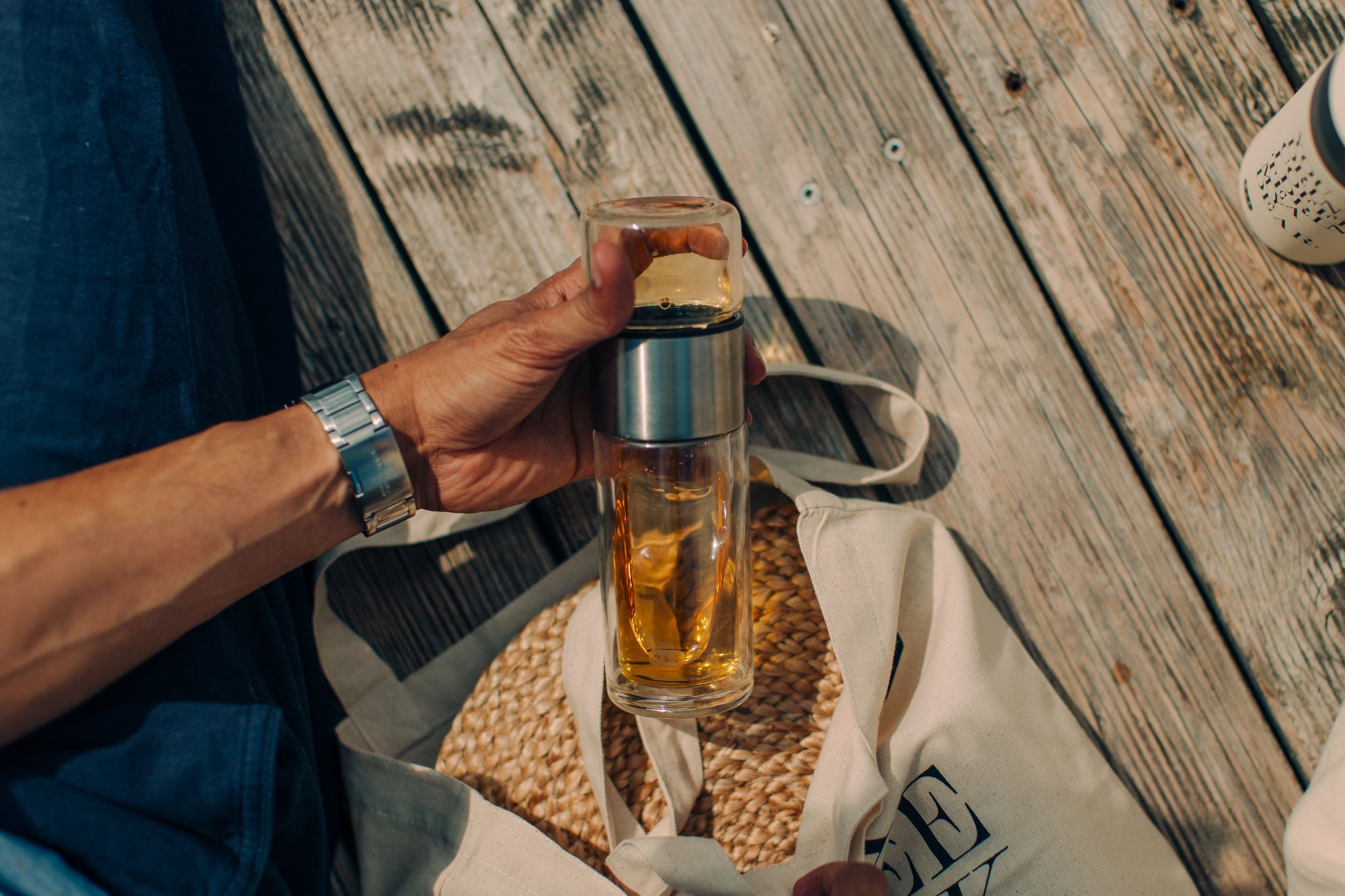 Person hält Glasflasche mit Metallband – Nomad Bottle – mit gelblicher Flüssigkeit, Fruchtstücken und Kräutern, sitzt auf Holzplanken neben beiger Tasche mit Text