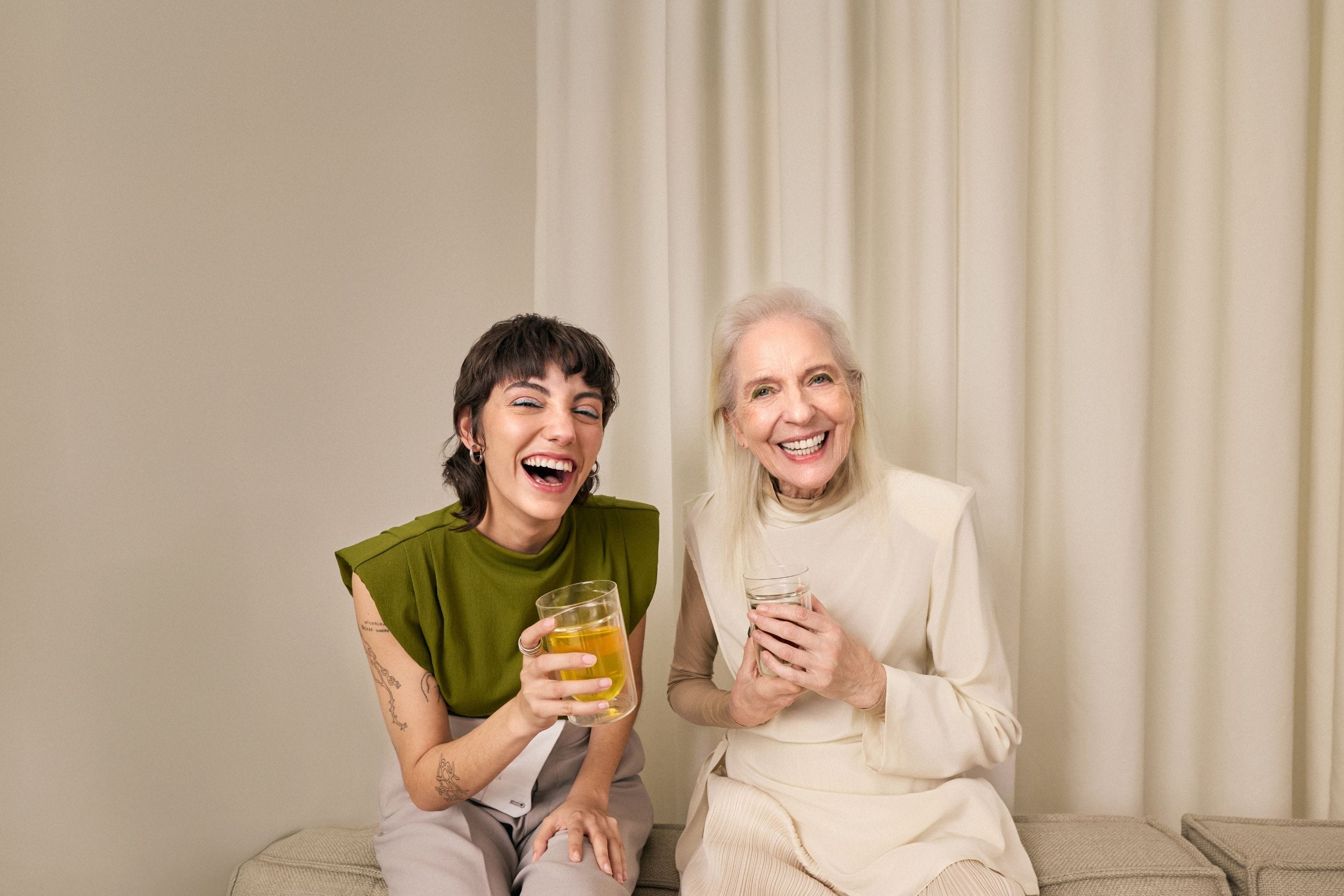 Zwei Personen sitzen nebeneinander auf heller Bank, halten jeweils ein Glas mit orange-gelbem bzw. pinkem Tee-Getränk, vor hellen Vorhängen