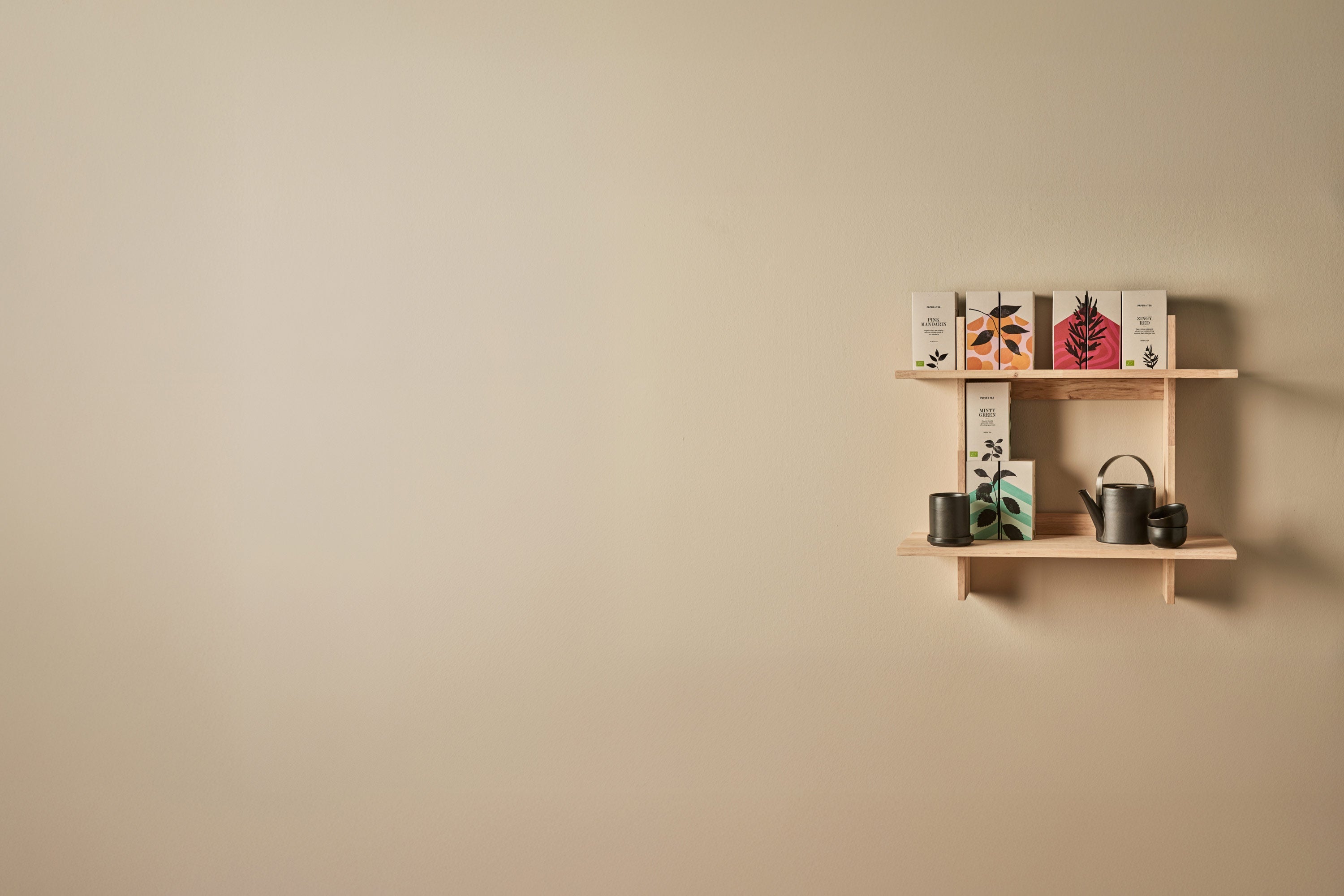 „Minimalistisches Holzregal an einer beigefarbenen Wand mit zwei Ebenen: oben stehen drei Teeboxen mit Pflanzenillustrationen, unten eine schwarze Teekanne und zwei Teebecher