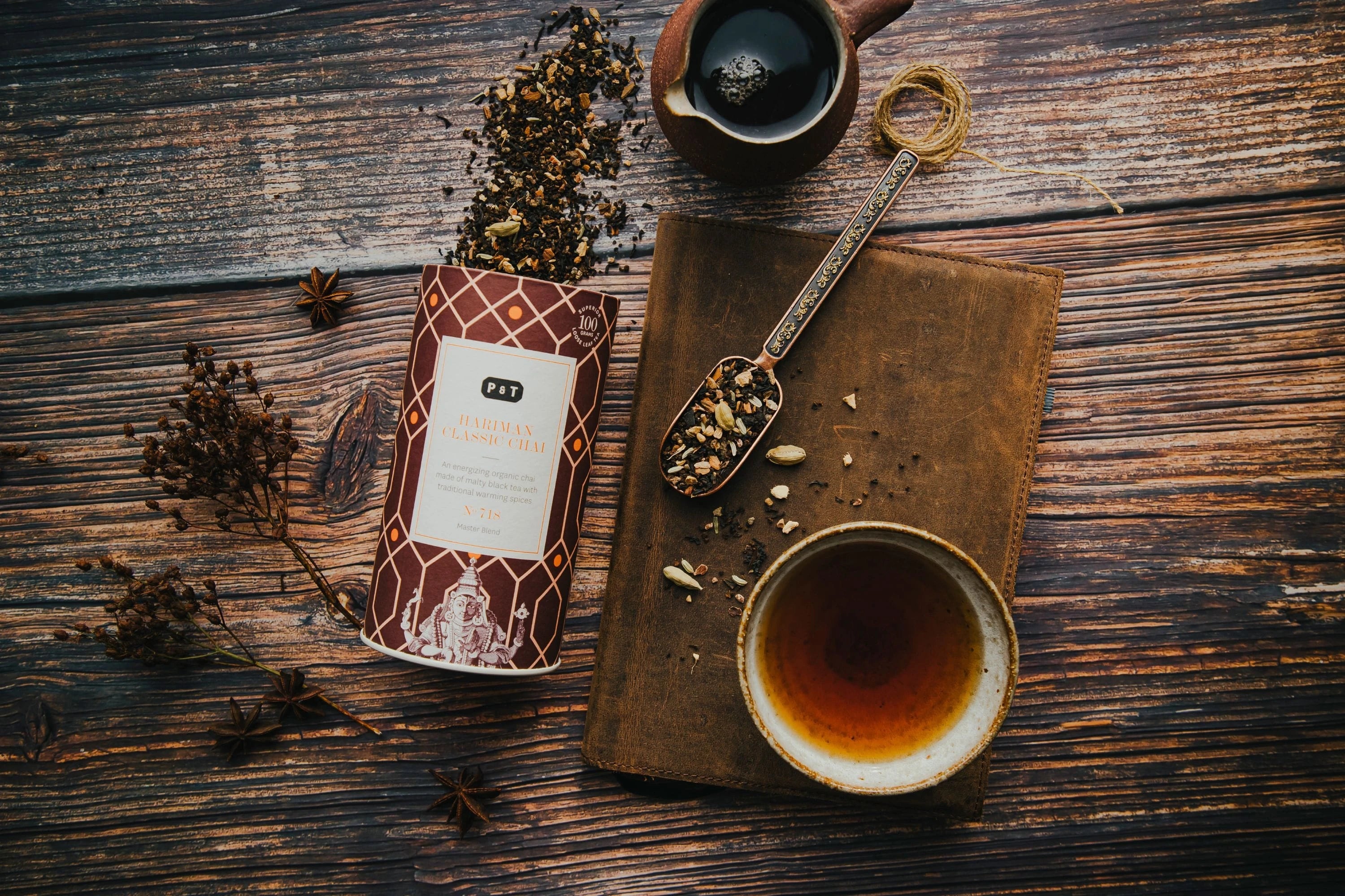 Rustikaler brauner Holztisch mit Teedose Hariman Classic Chai, altem Buch, Teetasse, Teelöffel mit losen Teeblättern, kleiner Teekanne und verstreuten Kräutern