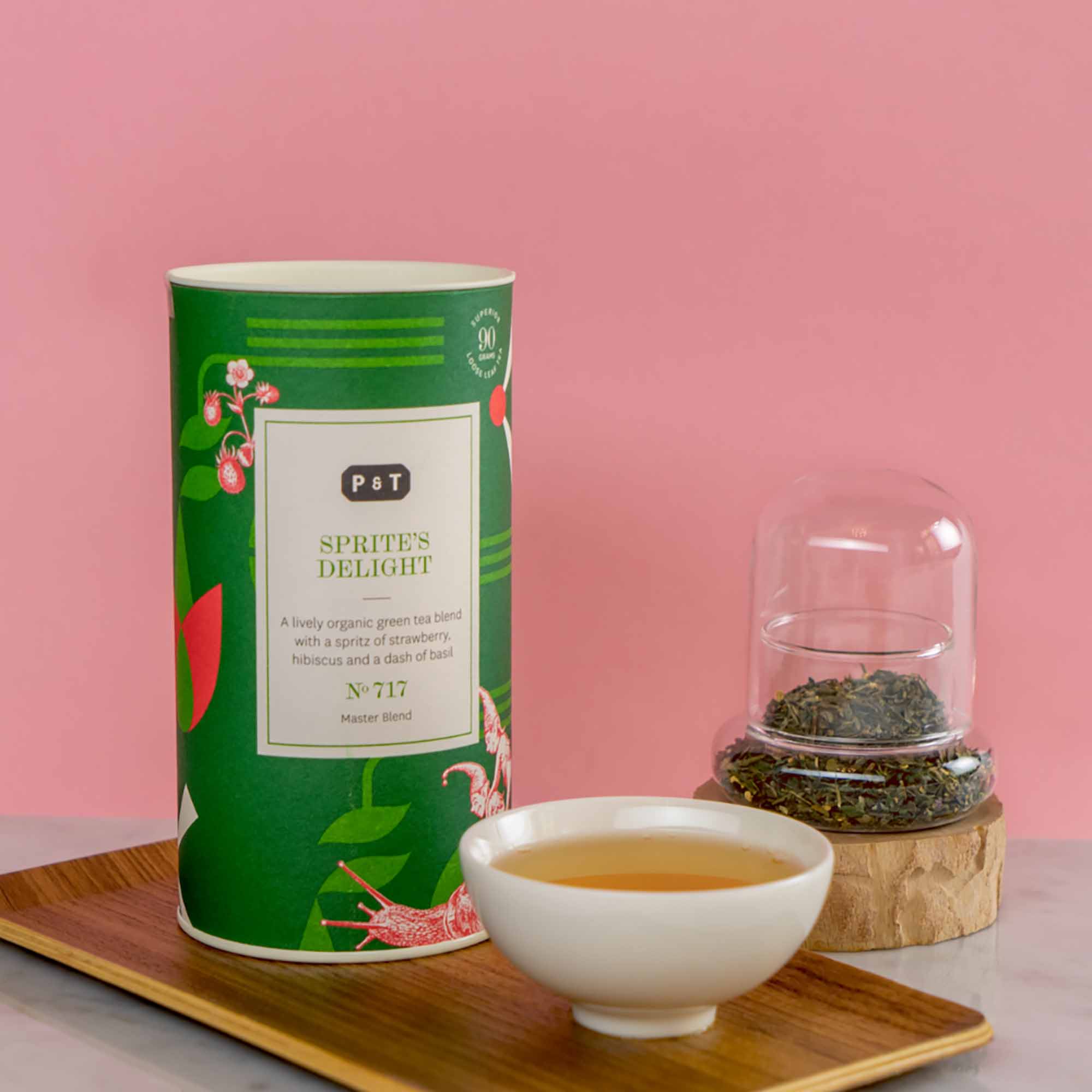 Grüne Teedose mit botanischen Illustrationen, neben Keramiktasse mit Tee und Glasglocke auf Holztablett vor rosa Hintergrund.