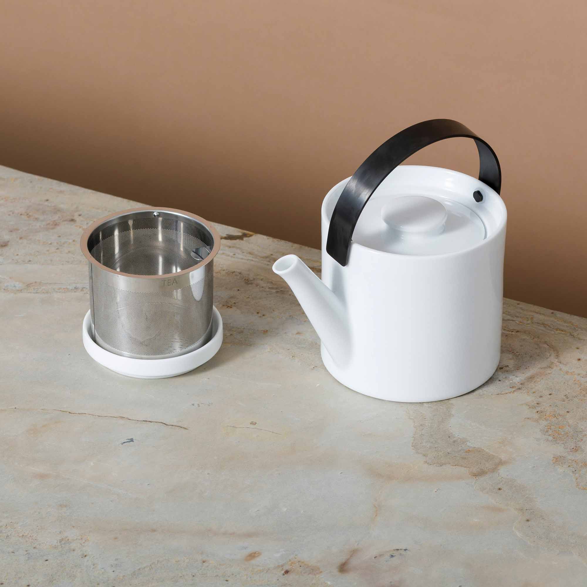 Weiße Keramik-Teekanne mit schwarzem Griff und feinem Teesieb auf Steinoberfläche vor braunem Hintergrund.