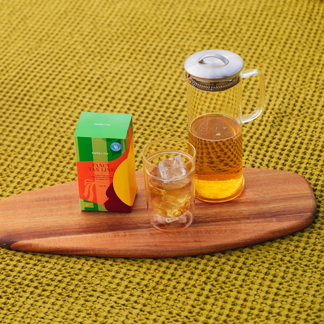Bunte PAPER & TEA Teebox mit Karaffe und doppelwandigem Glas Eistee auf Holzbrett vor gelbem Stoff.
