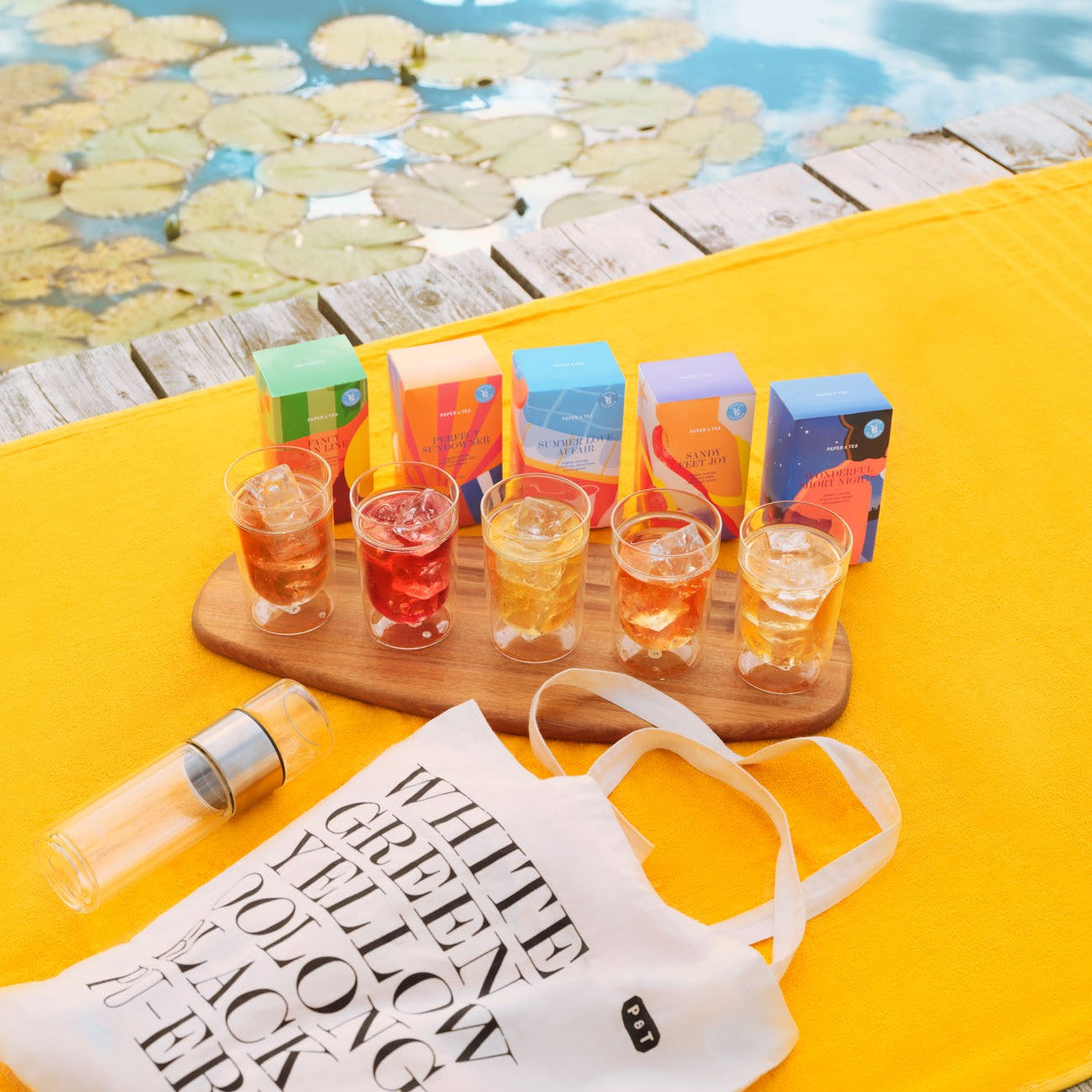 Fünf bunte Teeboxen mit eisgekühltem Tee auf Holzboden neben einem Pool und einer weißen Stofftasche mit Teesorten-Text.