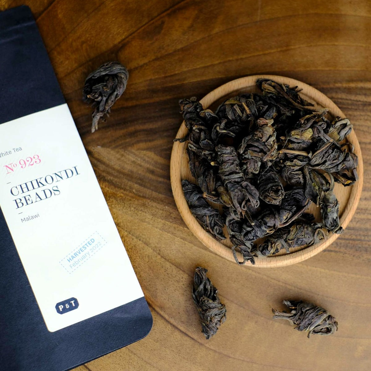 Holzschale mit weißen Teeblättern neben der Packung White Tea No 923 Chikondi Beads aus Malawi auf Holztisch.