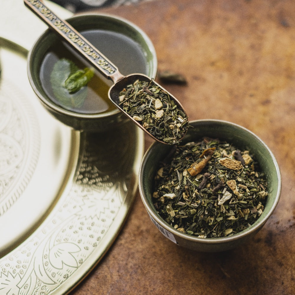 Grüner Tee in dekorativer Tasse mit Minzblatt, getrocknete Kräuter und kupferner Löffel auf goldenem Tablett