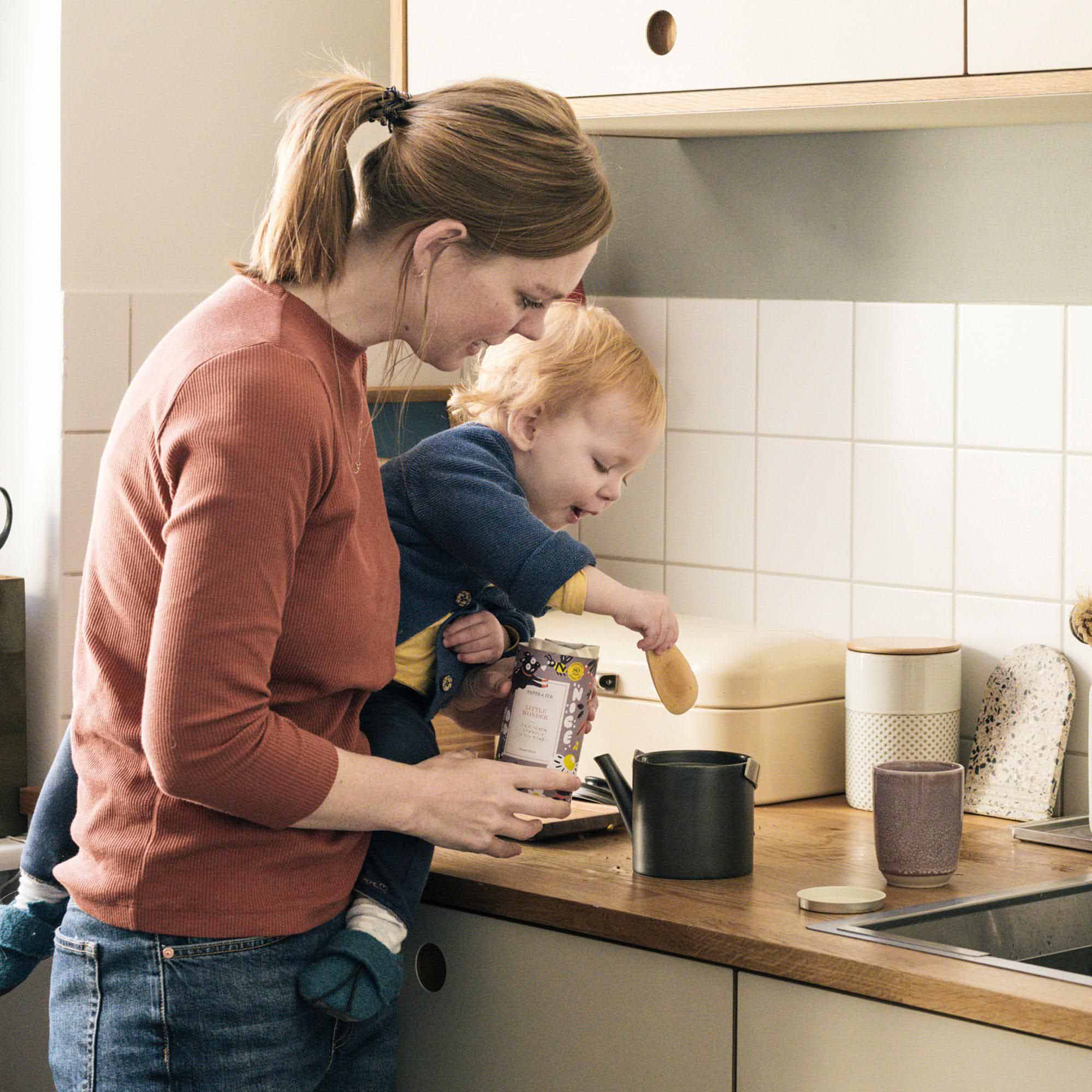 Frau in rostfarbenem Pullover hält Kind mit Holzlöffel in Küche mit Holzarbeitsplatte und cremefarbenen Schränken