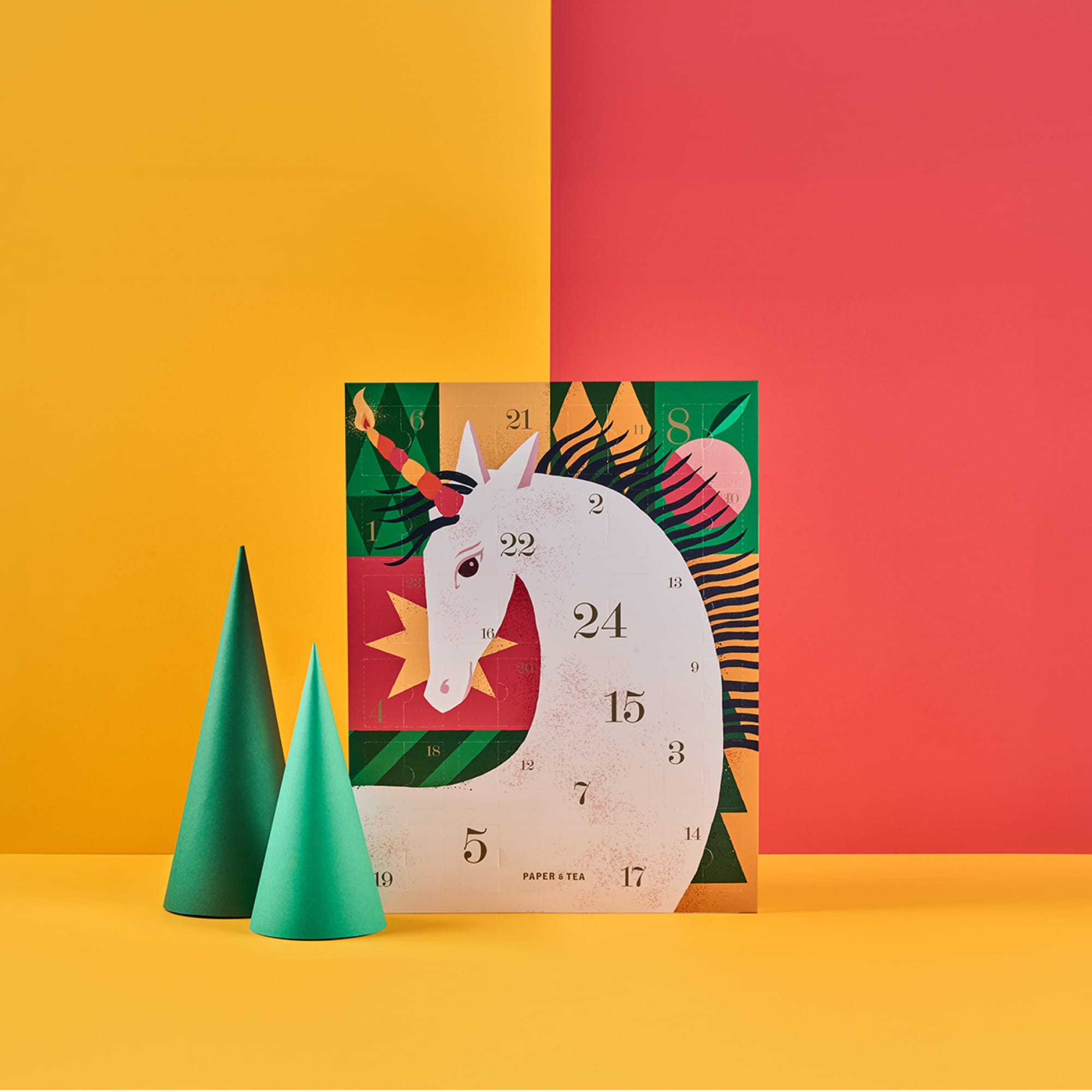 Bunter Einhorn-Adventskalender mit nummerierten Türen und grünen Papier-Weihnachtsbäumen vor gelb-rotem Hintergrund.