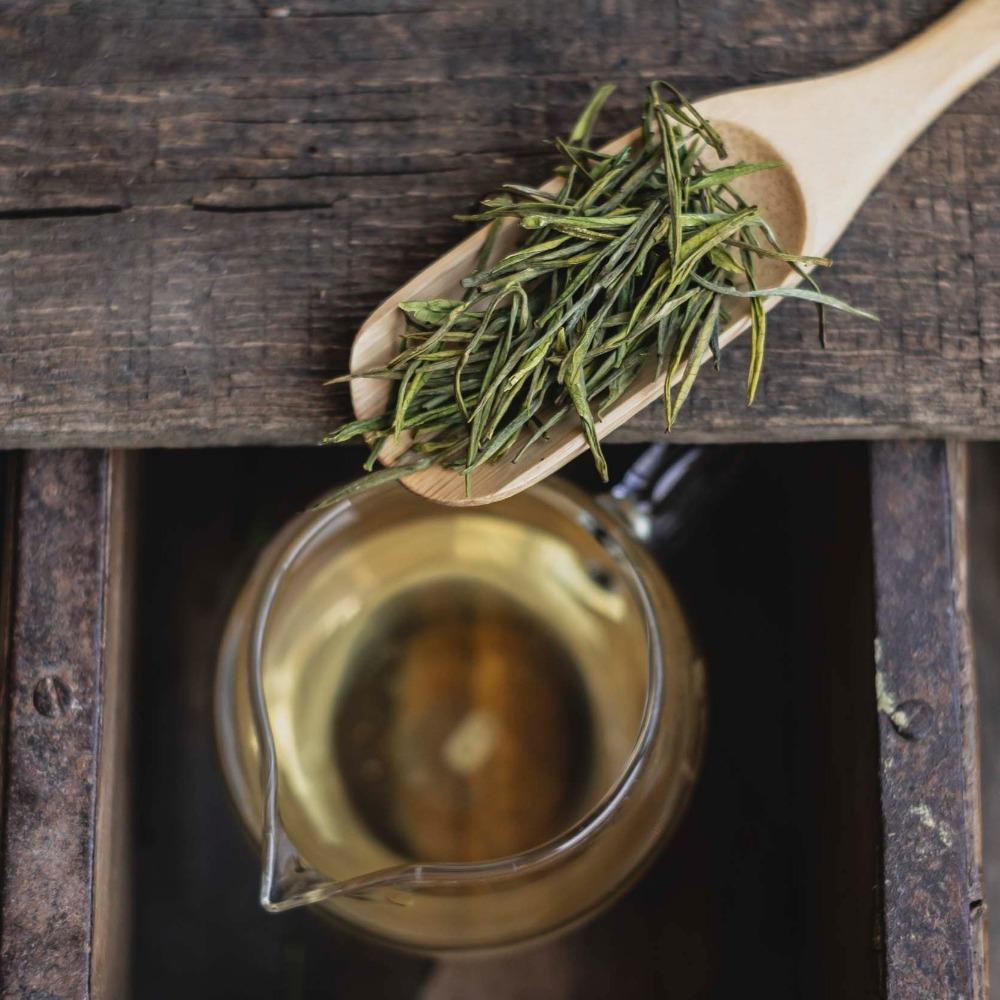 Holzlöffel mit getrockneten grünen Teeblättern neben Glas mit grünem Aufguss auf rustikalem Holztisch