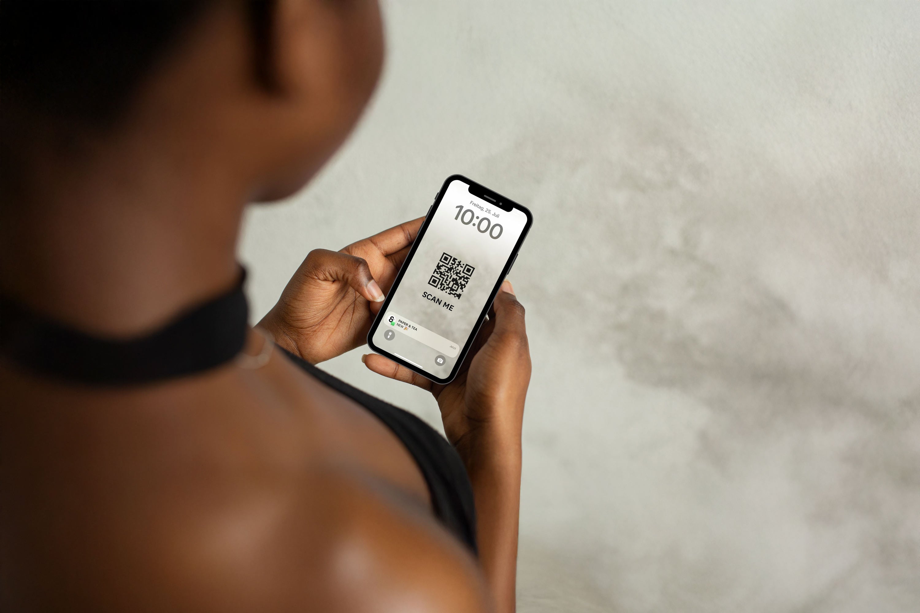 Person mit schwarzem Choker hält Smartphone mit QR-Code von Paper & Tea vor grauer Wand.