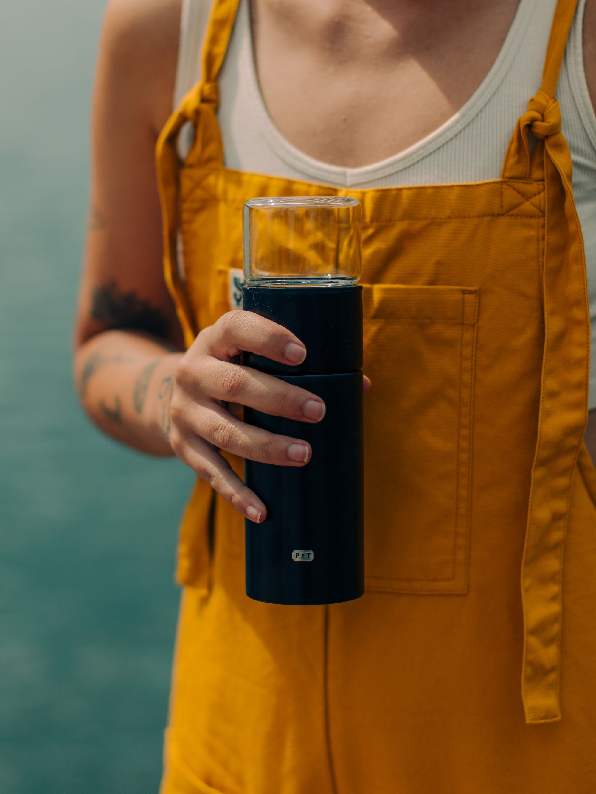 Person mit senffarbenem Latzkleid hält ein schwarzes zylindrisches Teegefäß mit P & T Etikett vor blauem Hintergrund.