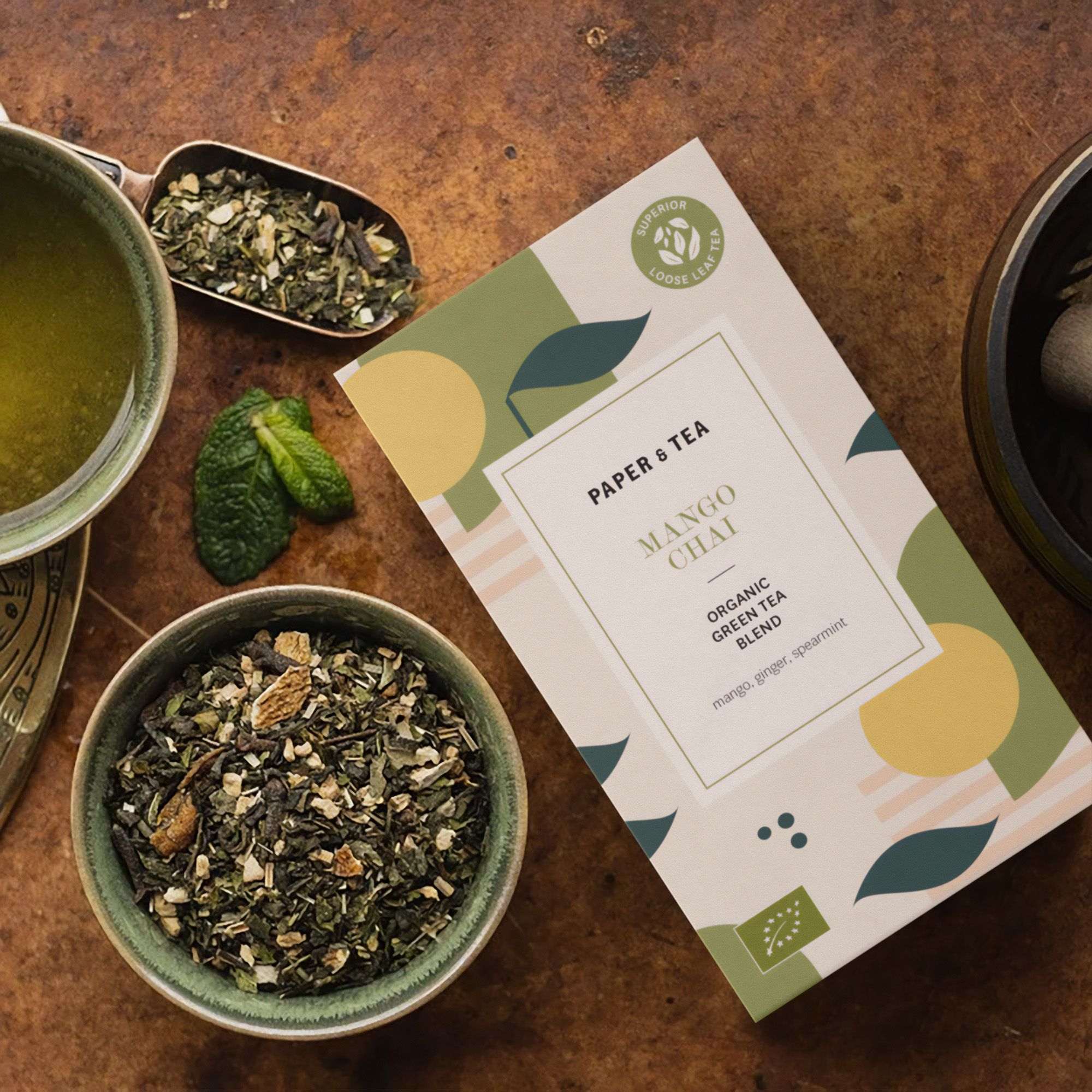 Moderne Verpackung für Mango Chai mit Bio-Grüntee-Mischung und Minze, daneben grüner Tee in Keramiktasse.