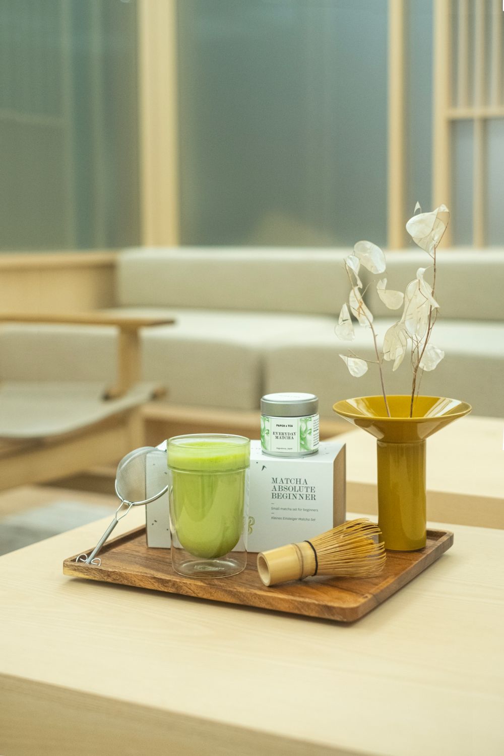 Holztablett mit grünem EVERYDAY MATCHA Tee im Glas, MATCHA ABSOLUTE BEGINNER Verpackung, Bambusbesen und gelber Vase.