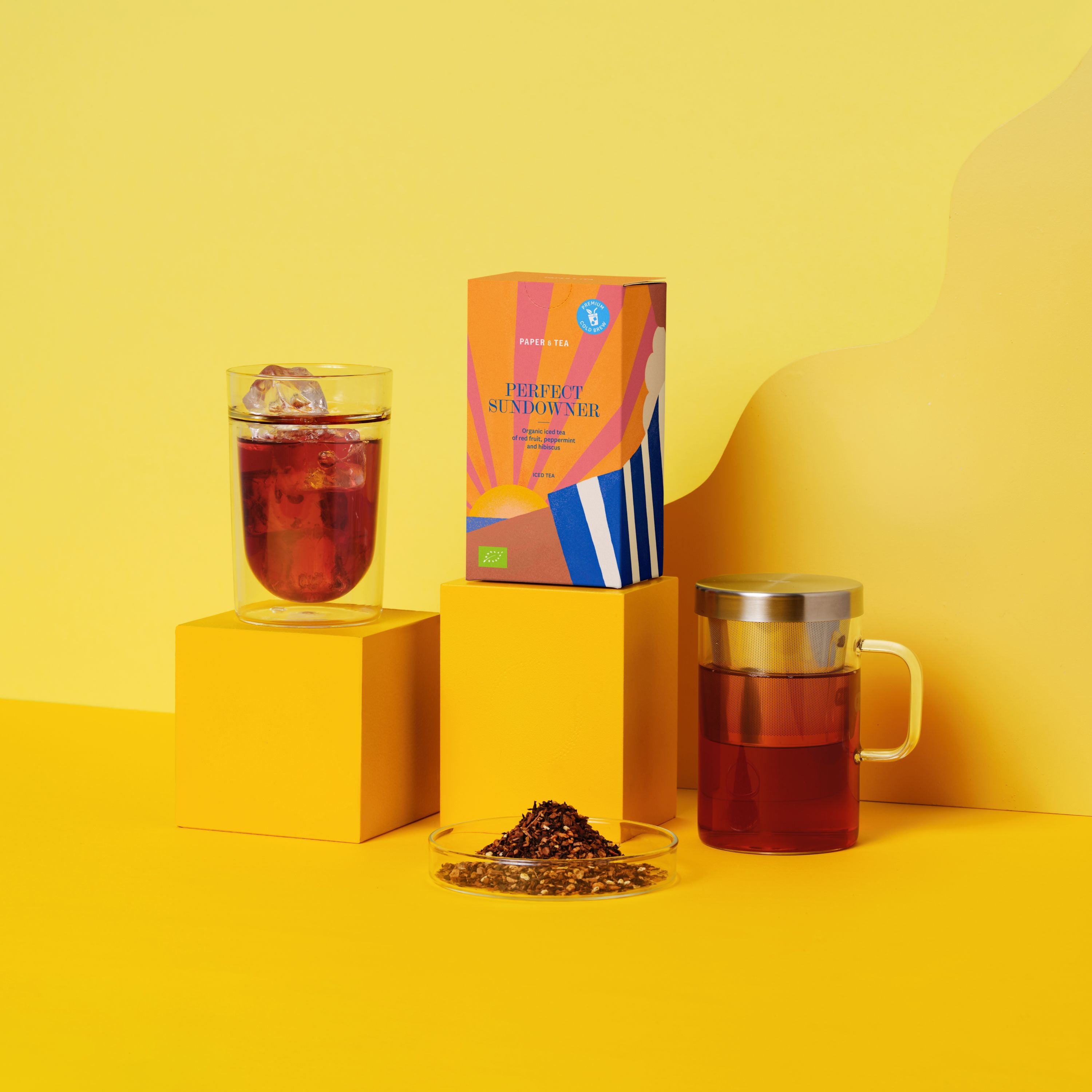 Bunte Verpackung des Paper & Tea Cold Brew Perfect Sundowner vor Glas mit Eis und Teekanne, gelber Hintergrund