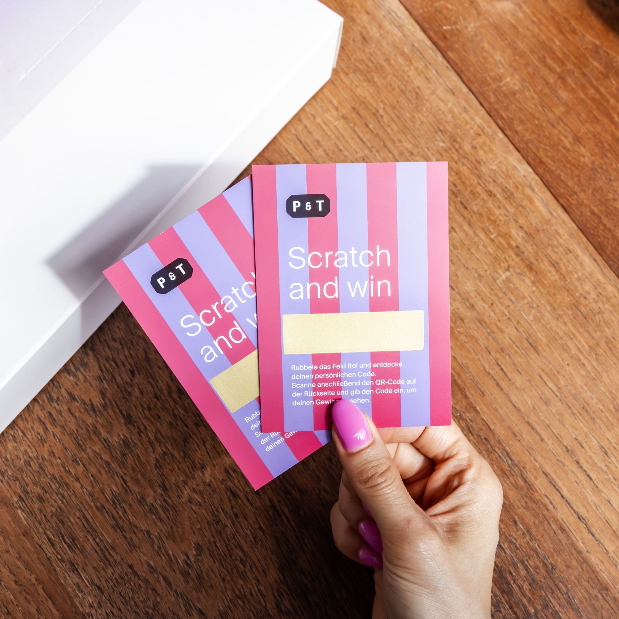 Hand mit pink lackierten Nägeln hält zwei P&T Rubbellose in Rot und Violett auf Holzuntergrund.