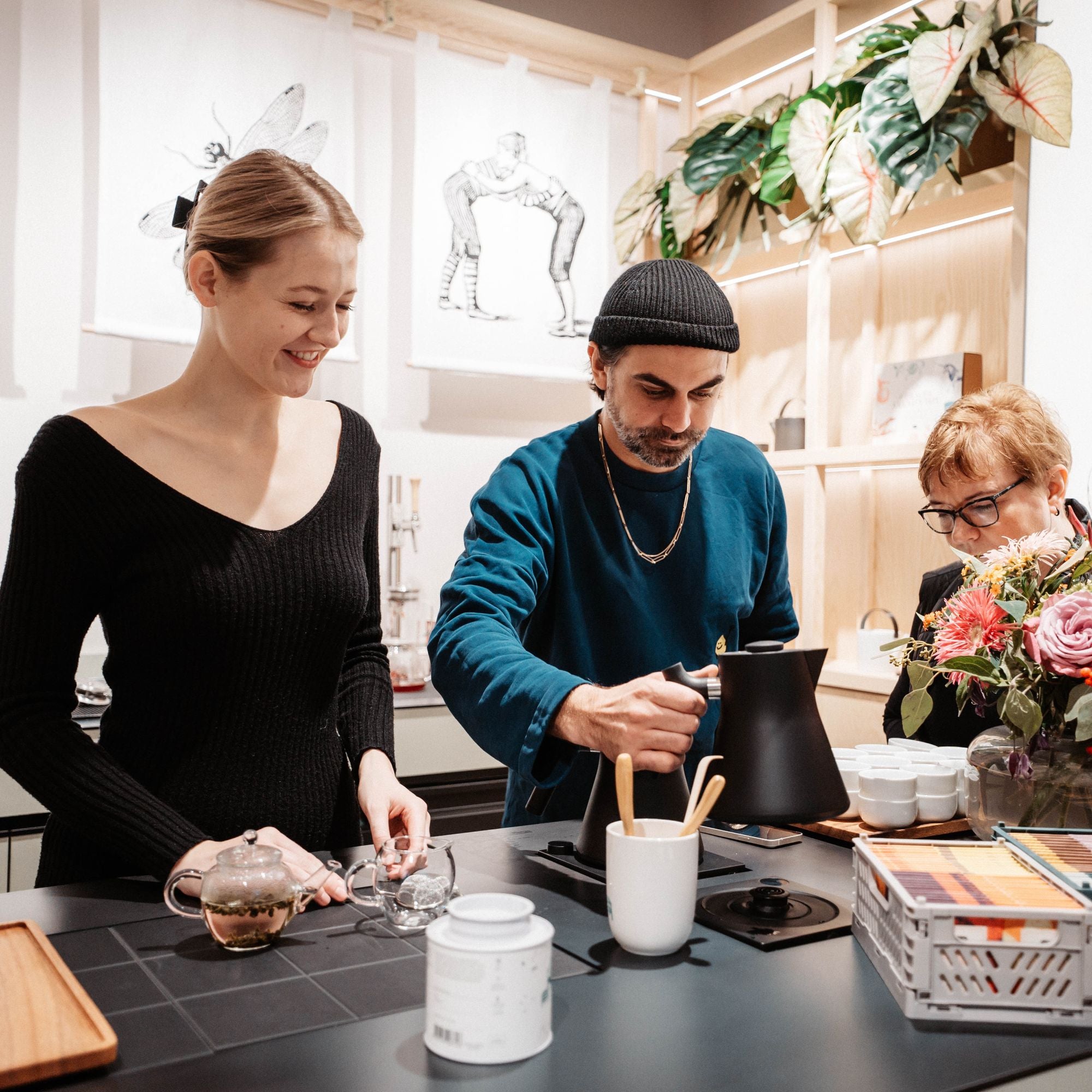 Junge Frau hält Glasteekanne, Mann gießt Wasser in Tasse, ältere Frau sitzt am Tisch.