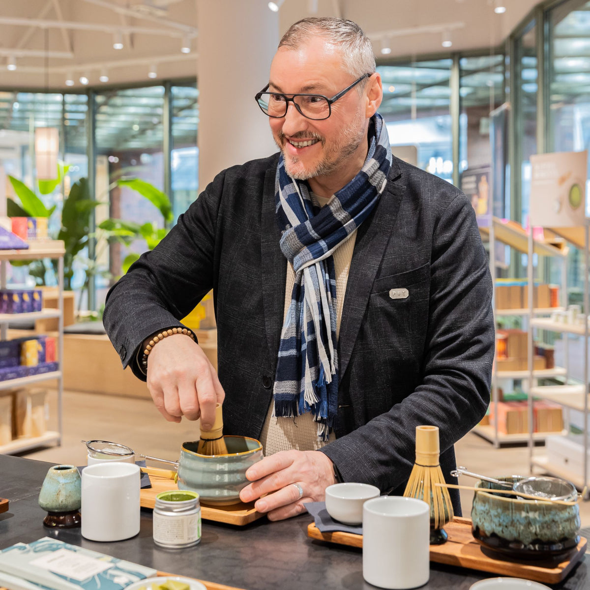 Mann mit Brille und blau-weiß gemustertem Schal bereitet Tee mit Bambusbesen in Keramikschale auf Holztablett zu.