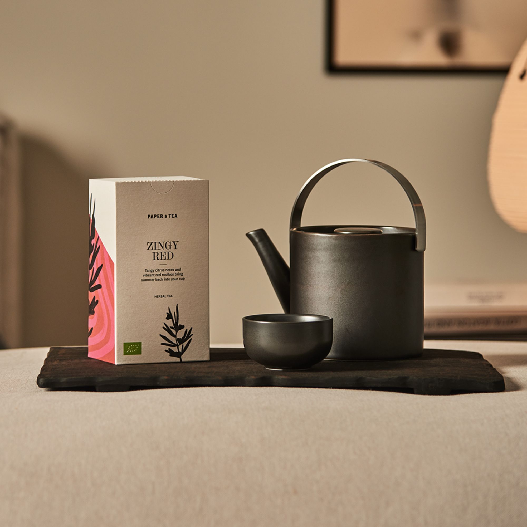 Minimalistisches Stillleben mit Paper & Tea Zingy Red Kräutertee, dunkler Teekanne und Becher auf Holztablett.