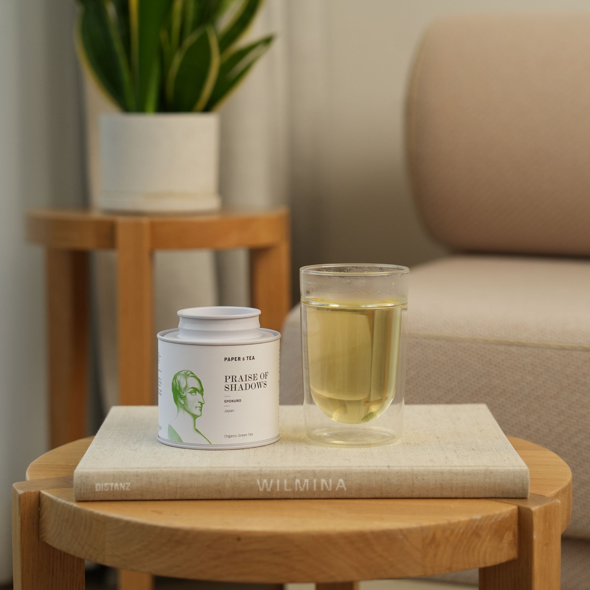 Weißer Tee-Behälter mit japanischem Bio-Gyokuro in einer klaren Tasse auf Buch DISTANZ WILMINA auf Holztisch.