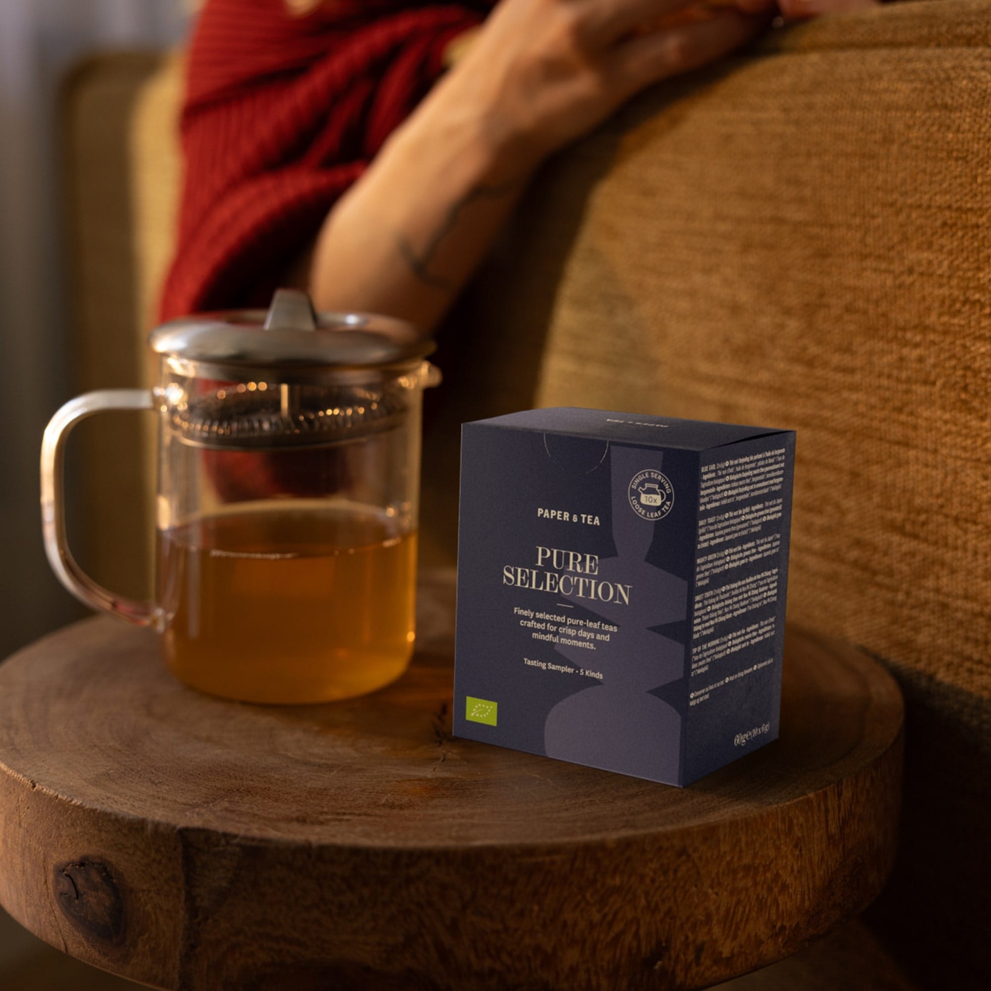 Blauer Tee Selection Probierset und klarer Teekrug auf Holztisch vor rotem Pullover.