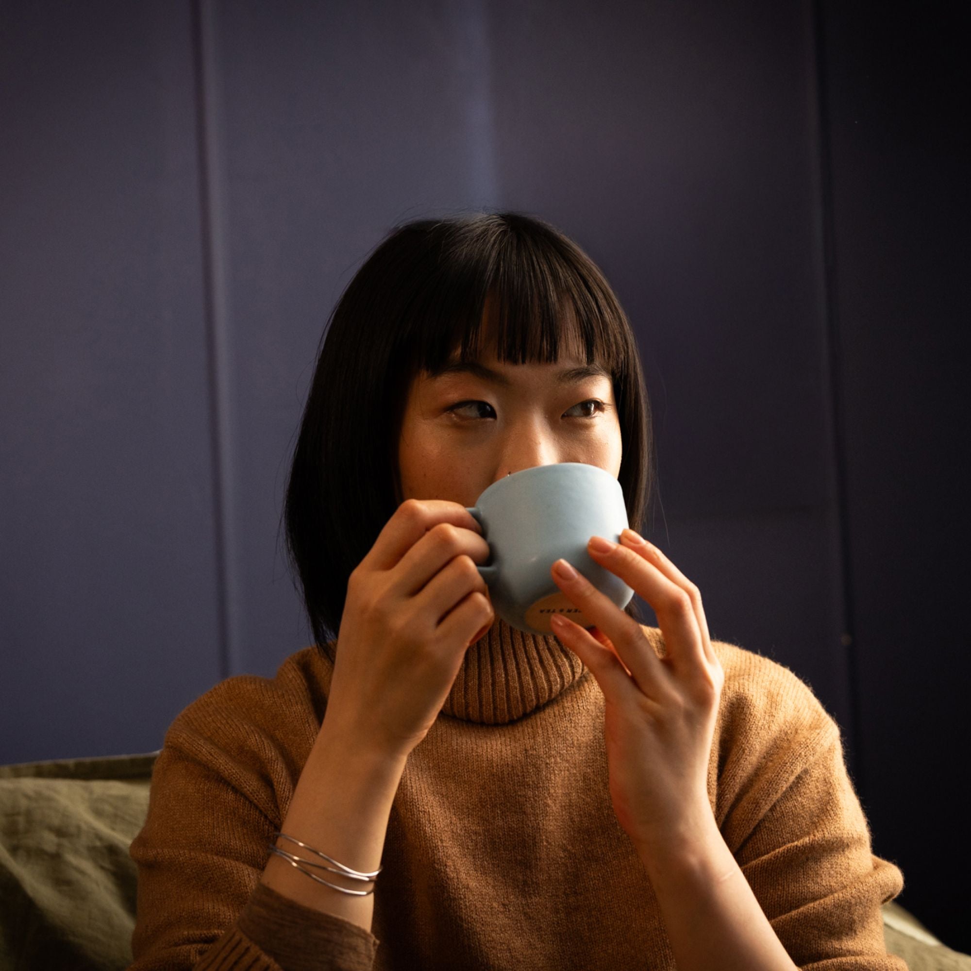Frau in braunem Rollkragenpullover trinkt Tee aus hellblauer Keramiktasse vor dunkelblauem Hintergrund.