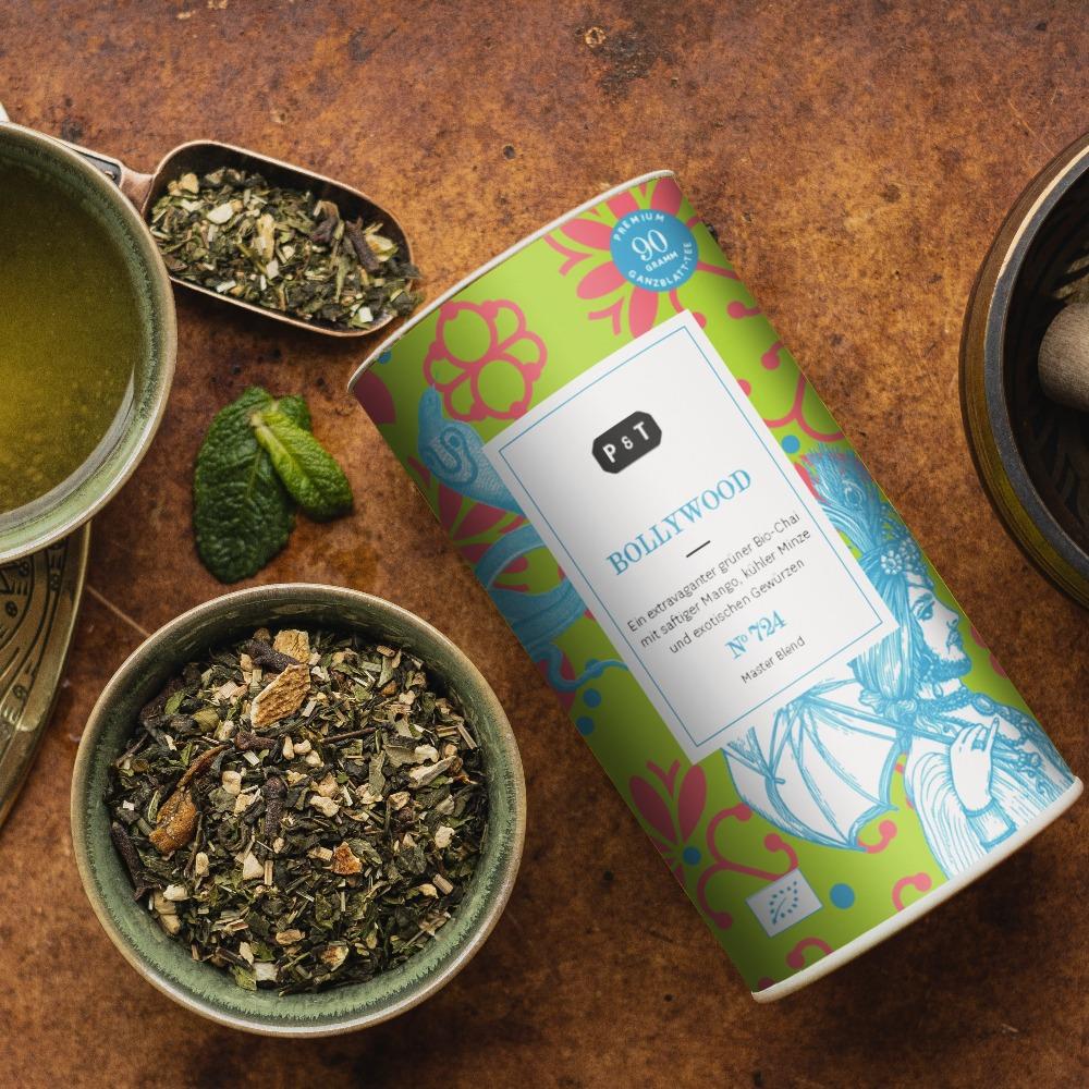 Grüner Bio-Chai 'BOLLYWOOD' in bunter Dose, umgeben von loser Teemischung, Minze und grünem Tee in Tasse.