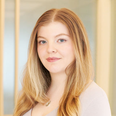 Junge Frau mit welligen hellbraunen Haaren und silberner Halskette vor hellem Hintergrund.