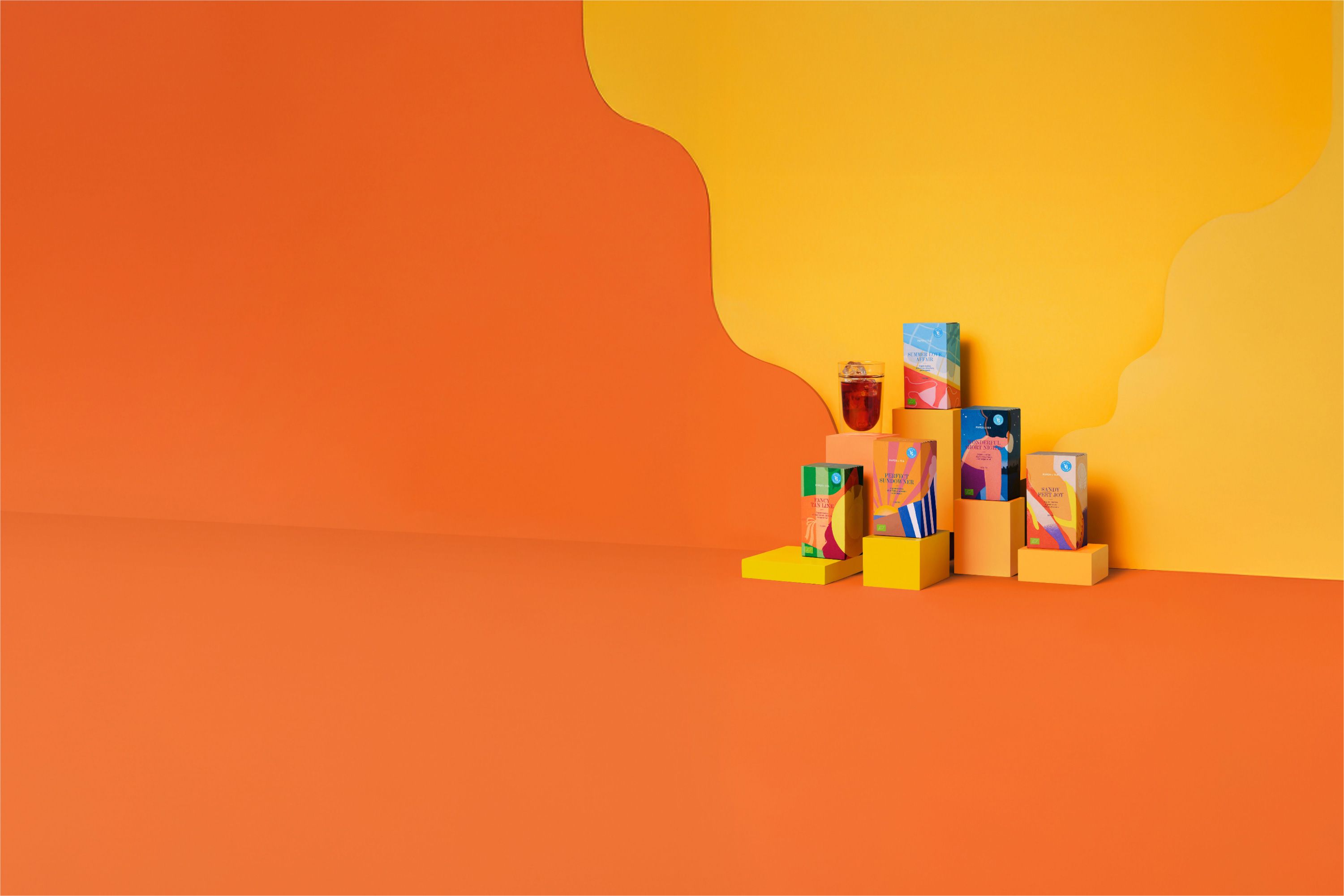 Fünf bunte Teepackungen in kreativen Designs auf Podesten neben einem Glas Eistee vor Sommerhintergrund.