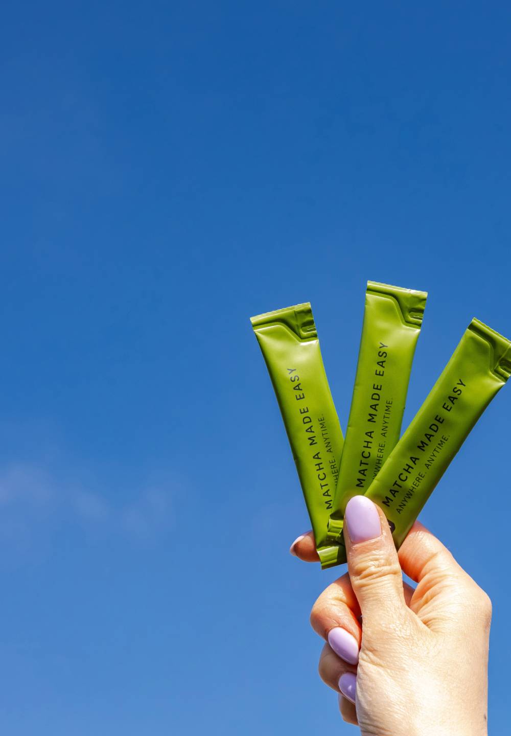 Hand mit lavendelfarbenen Nägeln hält drei schmale grüne P&T Matcha Made Easy-Päckchen vor klarem Himmel.