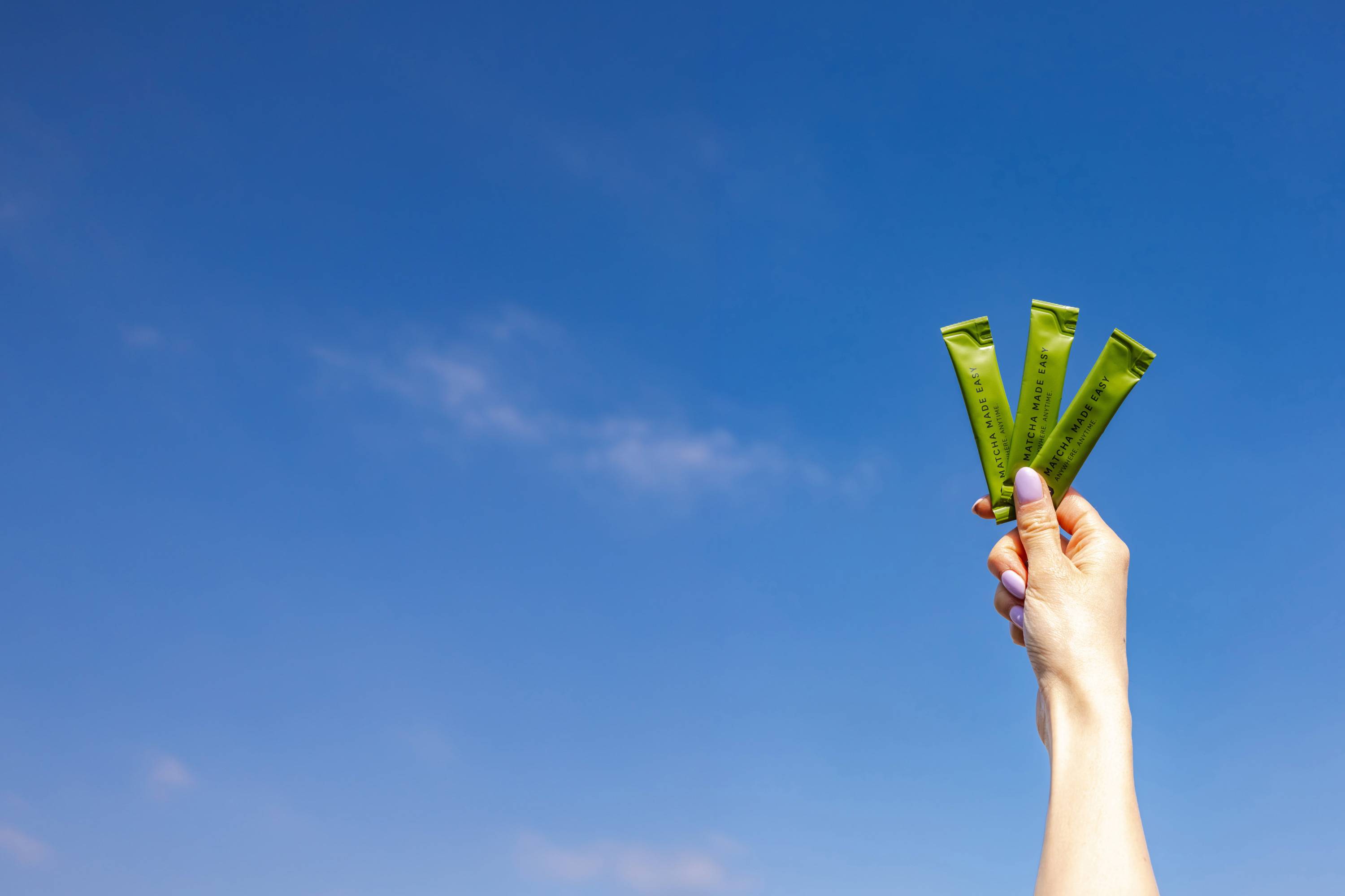 Hand mit violetten Nägeln hält drei grüne P&T Matcha Made Easy Sticks vor blauem Himmel mit Wolken.