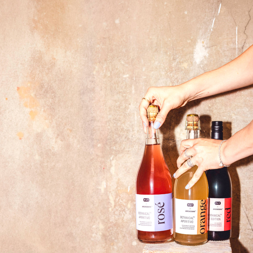 Drei Flaschen BOTANICAL APERITIVO mit rosé-, orange- und roten Etiketten, gehalten von Händen mit blau lackierten Nägeln vor beiger Wand.