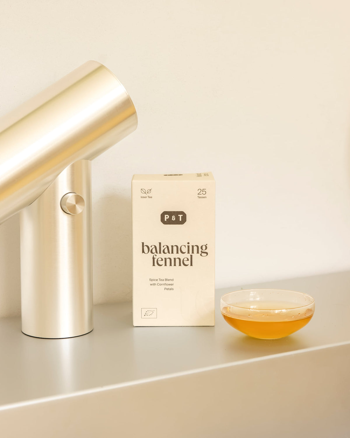 Beige rechteckige Verpackung von P&T Balancing Fennel Gewürz-Tee neben mattem Metallzylinder und Schale mit Teeaufguss.