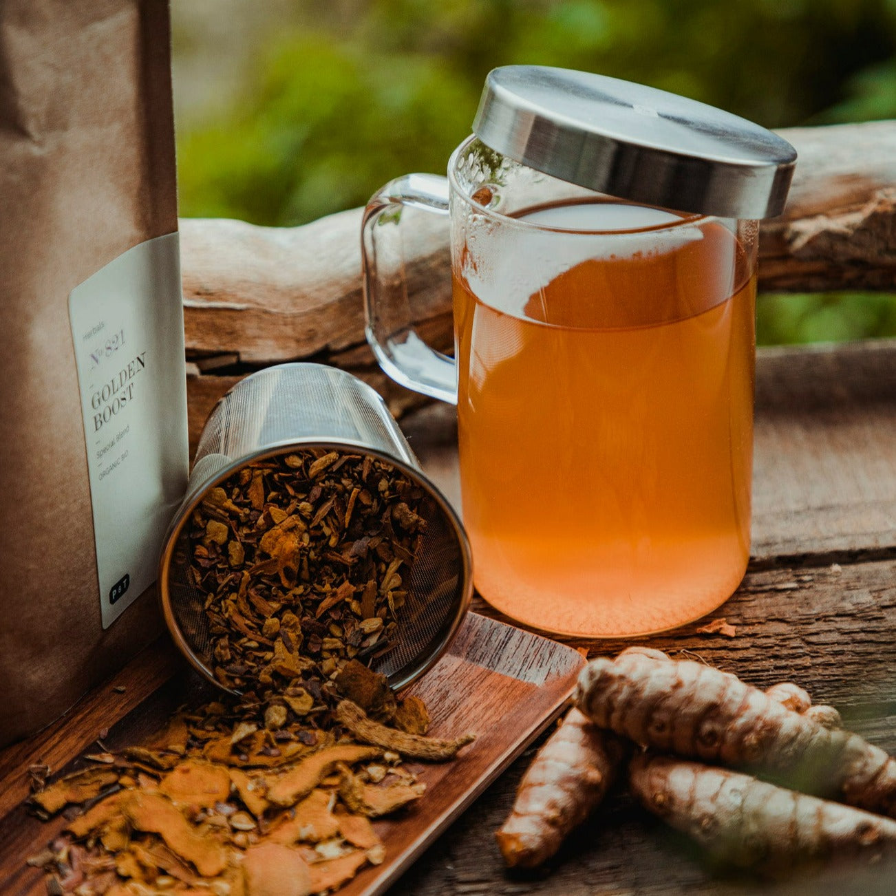 Glas mit goldenem Kräutertee, GOLDEN BOOST Bio-Tee in brauner Verpackung und Kurkuma-Tee auf Holztablett.