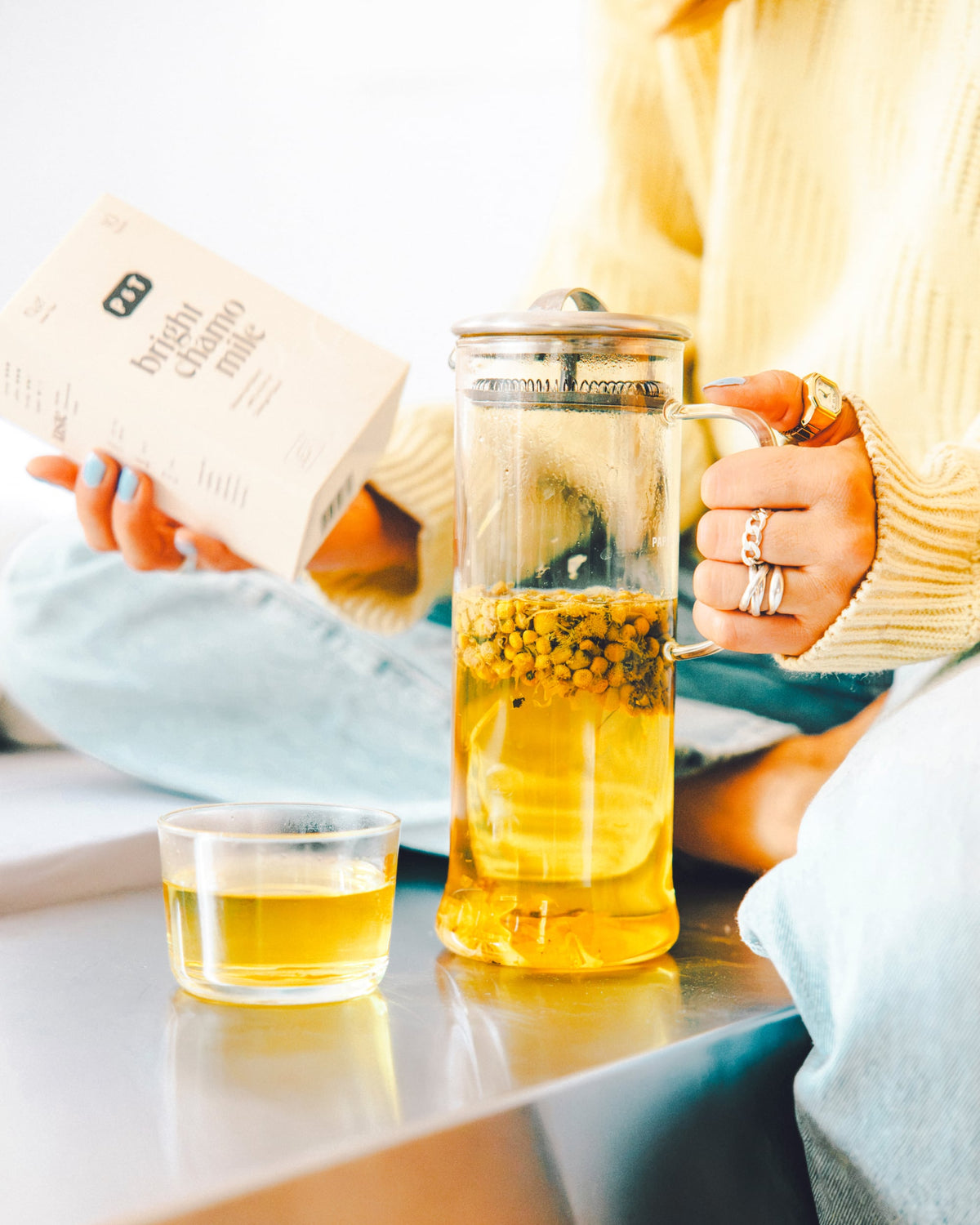 Person in Jeans und cremefarbenem Pullover hält P&T Teebox 'Bright Chamomile' und Glas-French-Press.