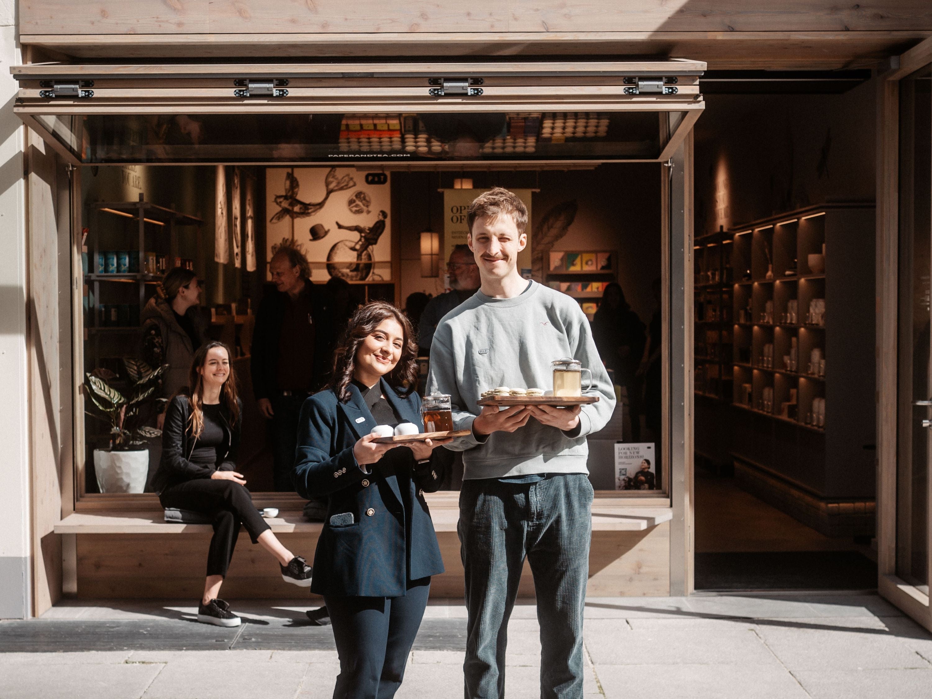 Junge Gastgeber im Teeladen präsentieren Holztabletts mit Teekannen und Tassen, entspannte Atmosphäre.