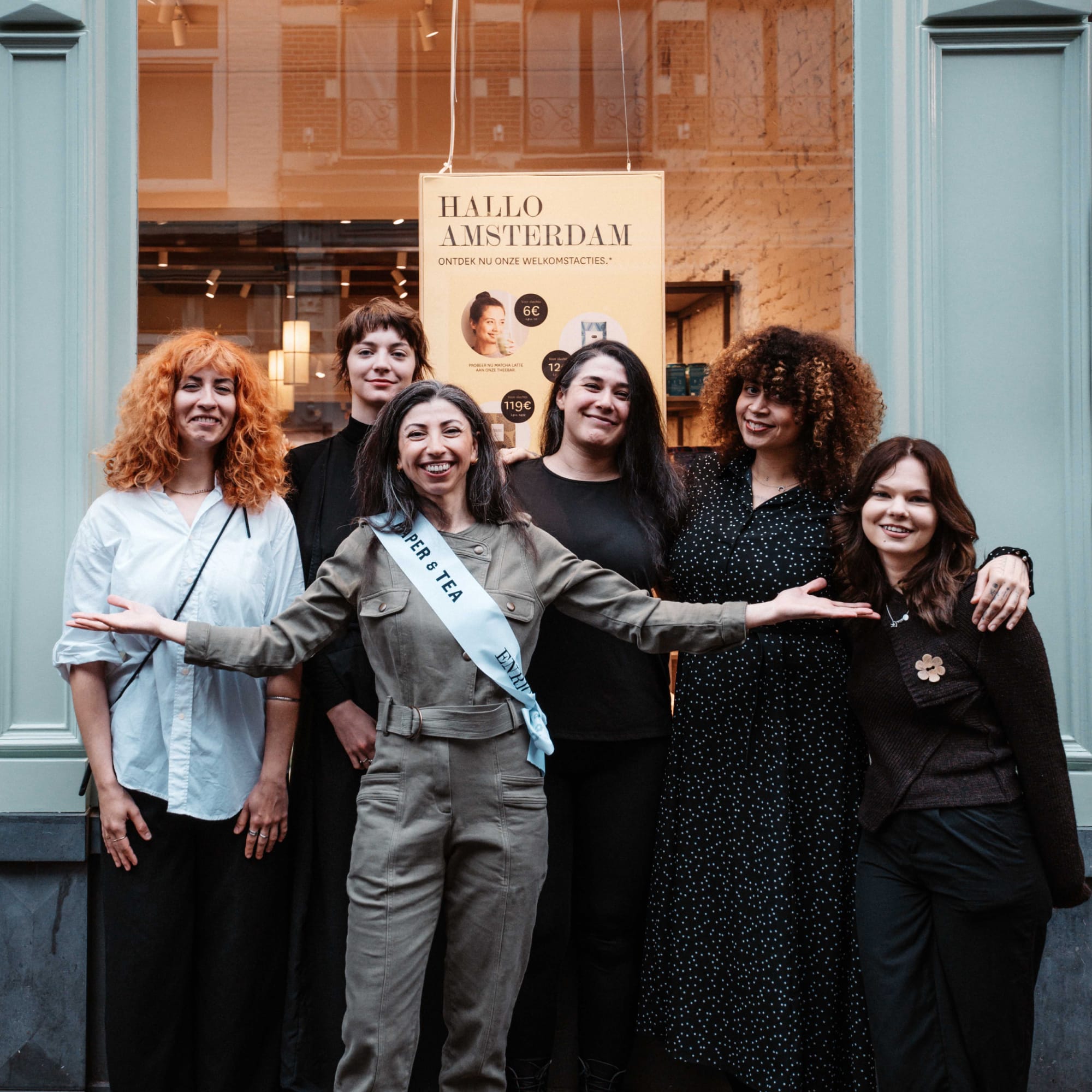 Sechs diverse Frauen vor der türkisfarbenen Tür von Paper & Tea Enri in Amsterdam, eine trägt eine Willkommensschärpe. Sie lächeln vor einem Poster für Matcha Latte Angebote.