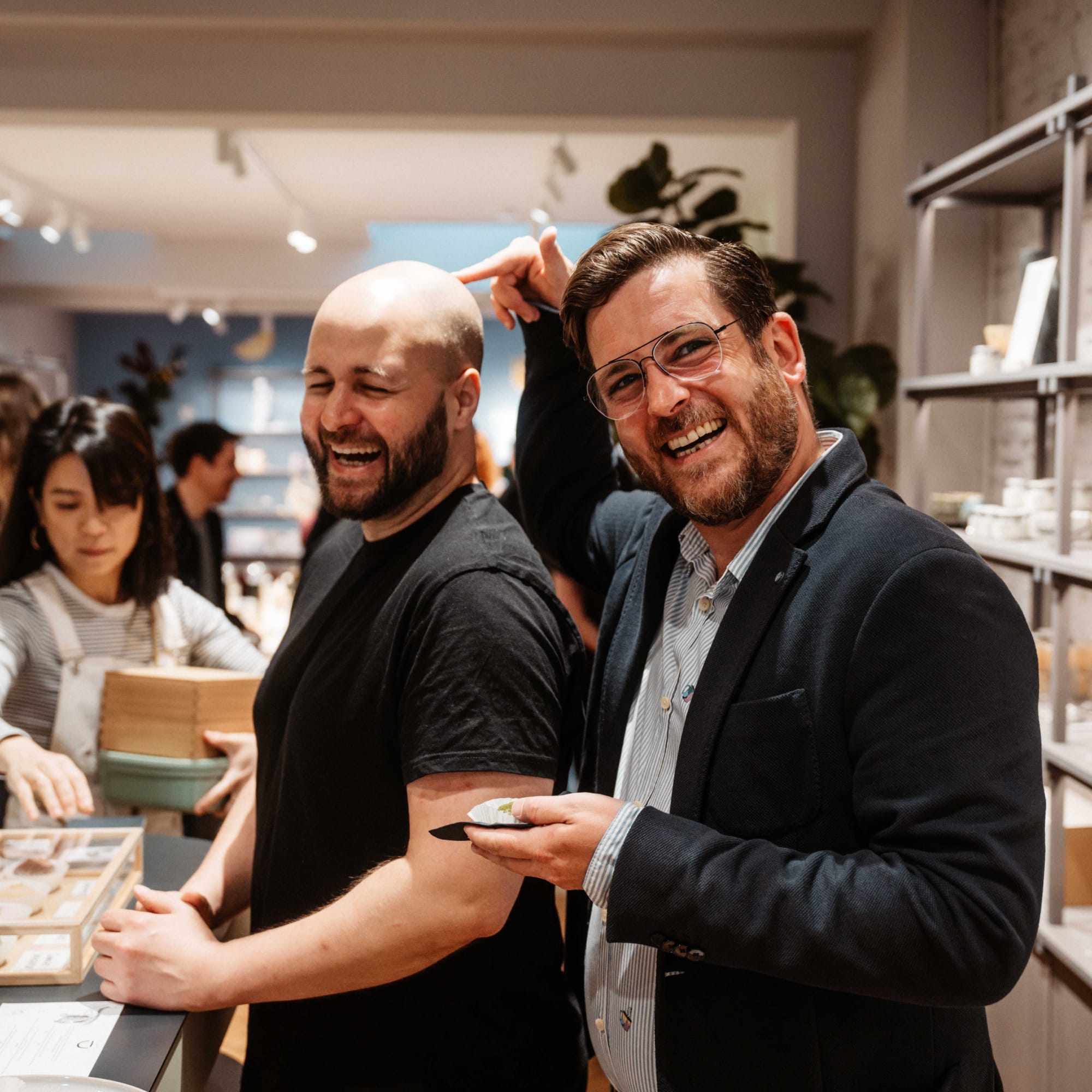 Zwei Männer lachen in einem modernen Teegeschäft; im Hintergrund ordnet eine Frau Produkte.