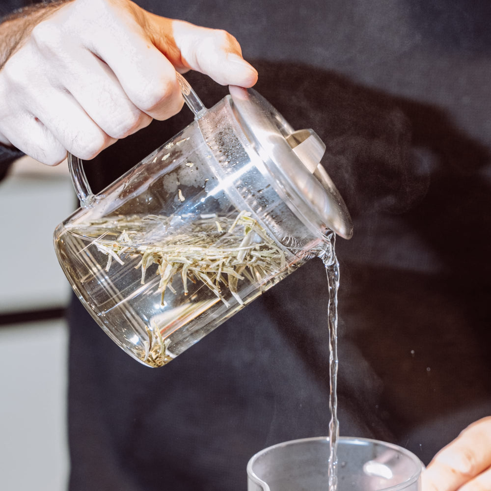 Hand gießt dampfenden Tee mit Teeblättern aus Glaskanne in Glastasse.