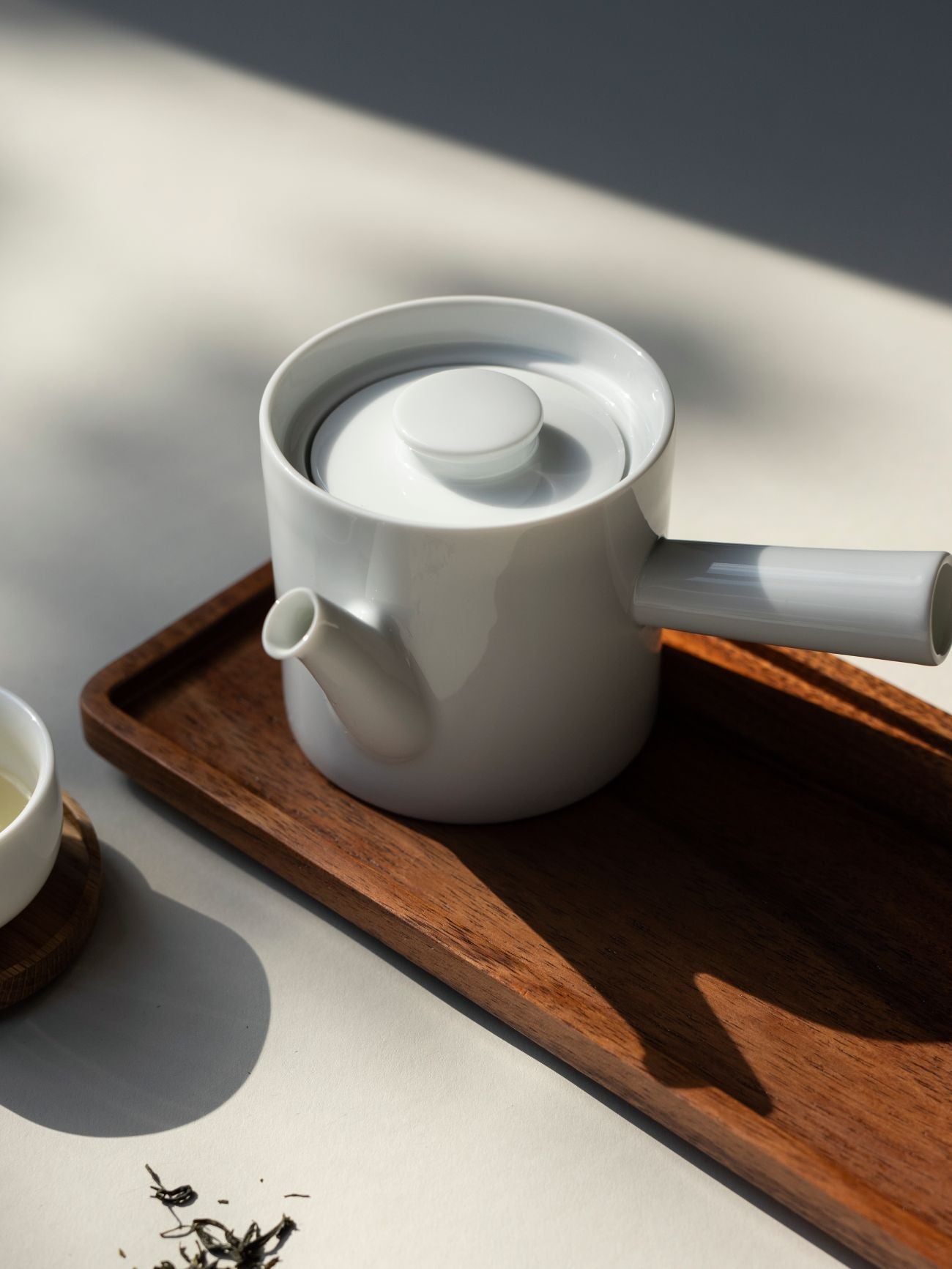 Weißer Keramik-Teekessel mit langem Griff auf Holztablett, umgeben von losen Teeblättern und Tasse auf Untersetzer.