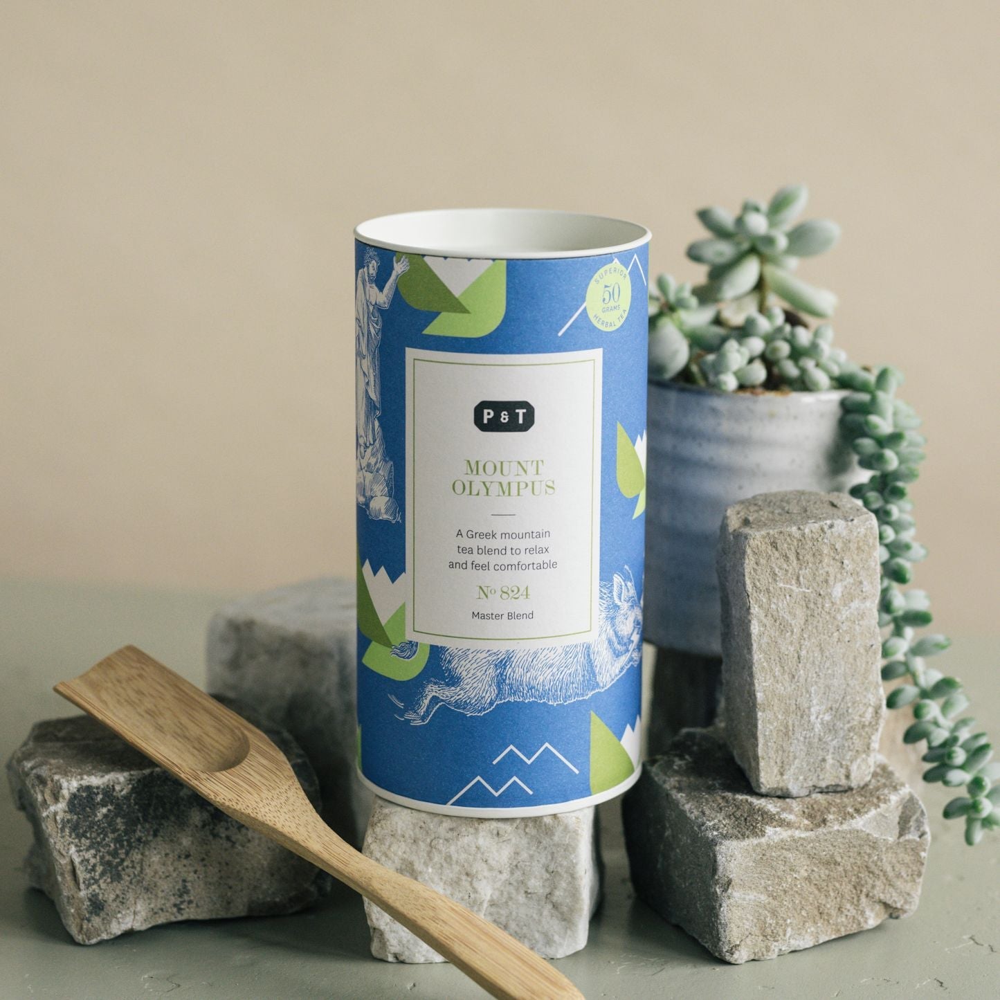 Zylindrische Teedose Mount Olympus mit blauem Etikett, umgeben von Natursteinen, Holzlöffel und Sukkulente.