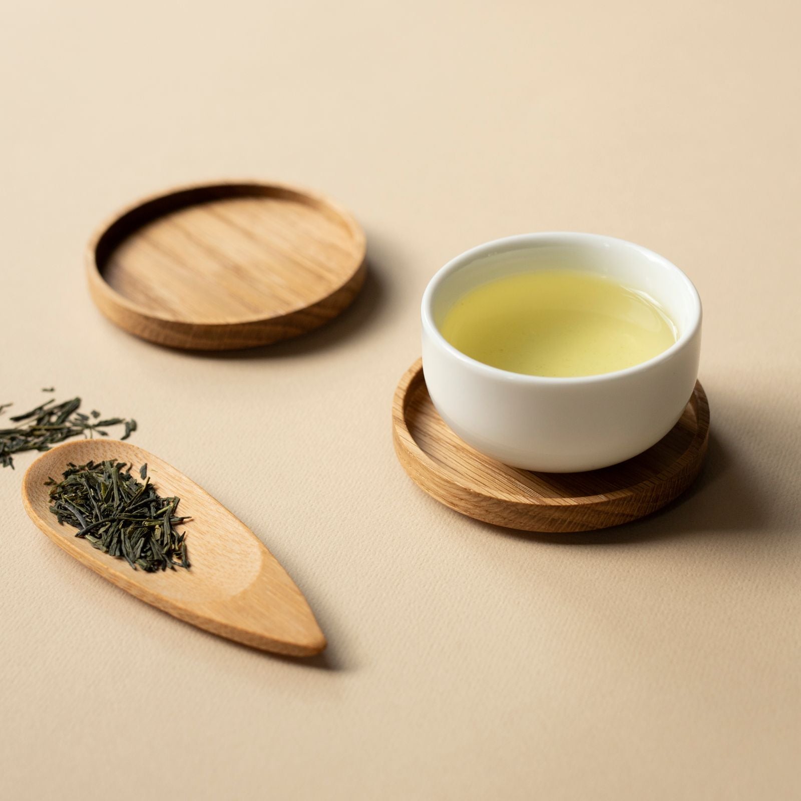 Weißer Keramikbecher mit hellgrünem Tee auf rundem Holzuntersetzer, daneben Holzlöffel mit grünen Teeblättern.