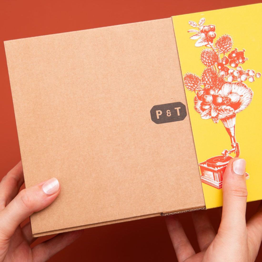 Zwei Hände halten eine Papierschachtel mit P & T-Label und gelbem Booklet mit Blumen vor rotem Hintergrund.