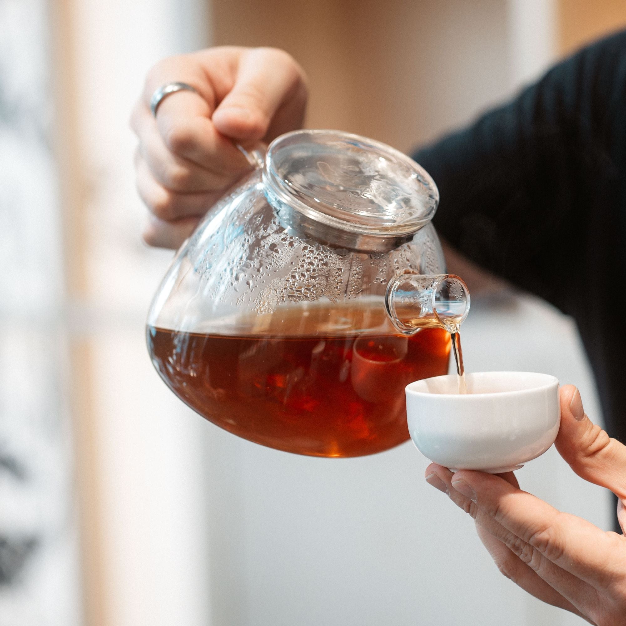Eine Hand gießt frisch gebrühten Tee aus einem Glas-Teekessel in eine weiße Keramiktasse.