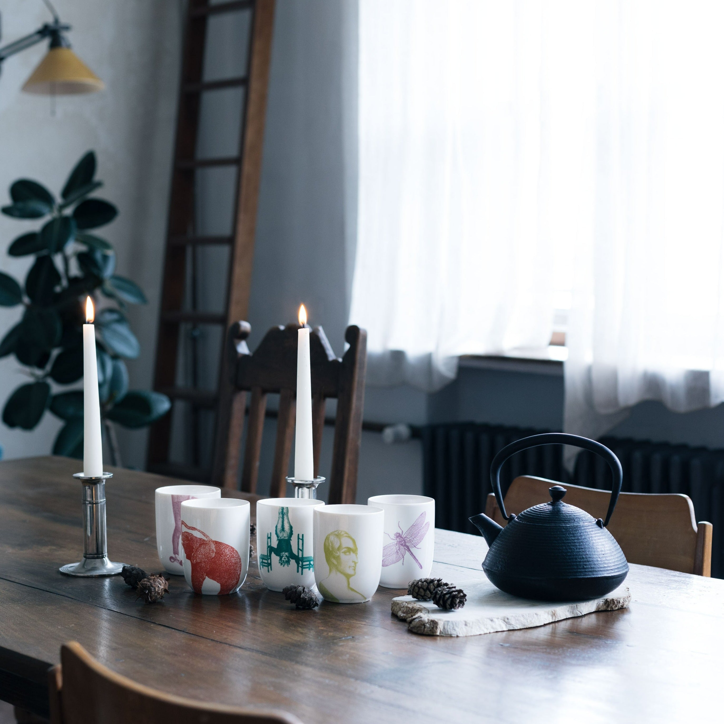 Rustikaler Holztisch mit weißen Keramiktassen, gusseisernem Teekessel und brennenden Kerzen.