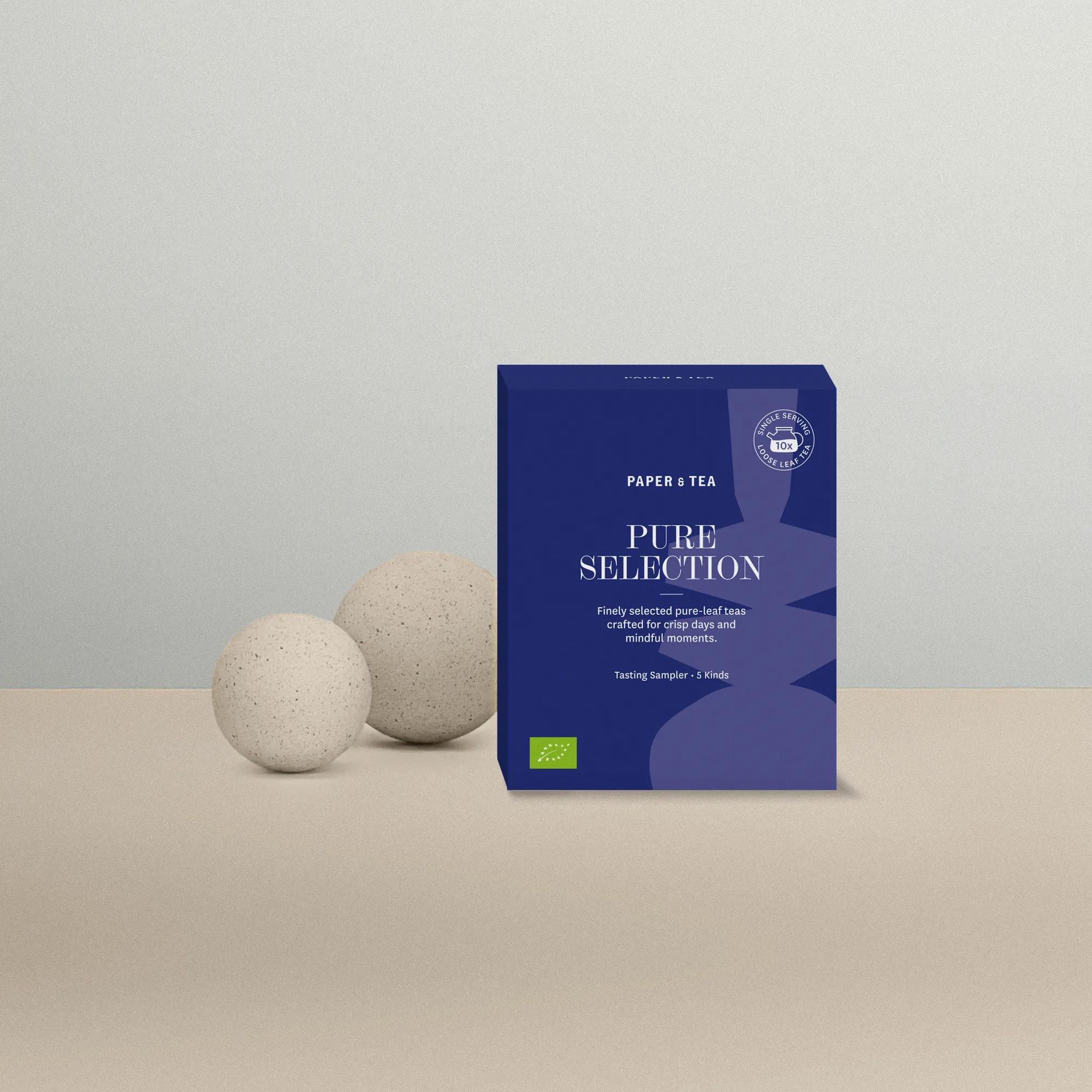 Blaues Paket mit losem Tee Pure Selection von Paper & Tea, vor grauem Hintergrund und Steinkugeln.