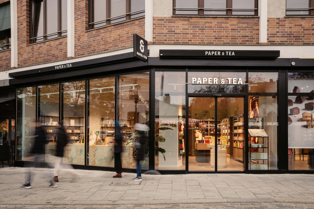 Modernes PAPER & TEA Geschäft mit großen Glasfronten und schwarzen Schildern an einem Backsteingebäude.