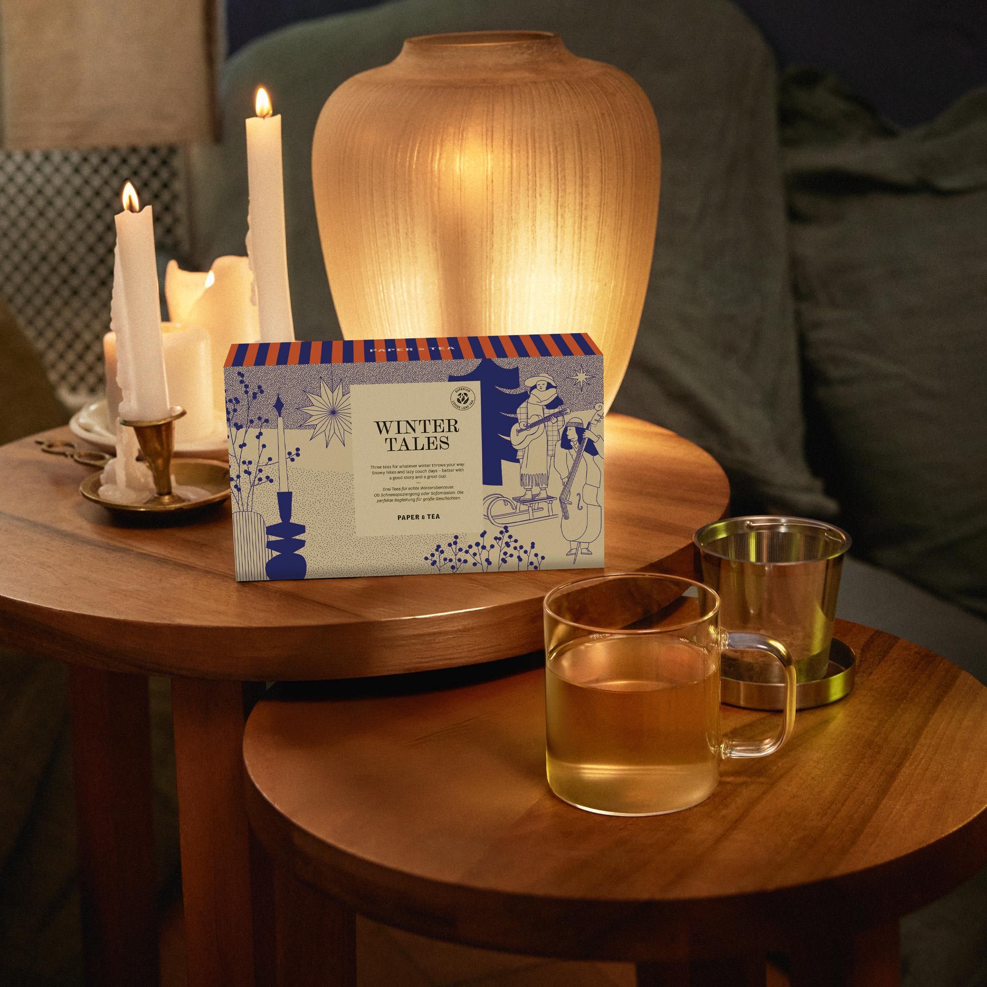 WINTER TALES Teebox auf Holztisch, mit Glasbecher Tee, Teeei und Kerzen im warmen Lampenlicht.