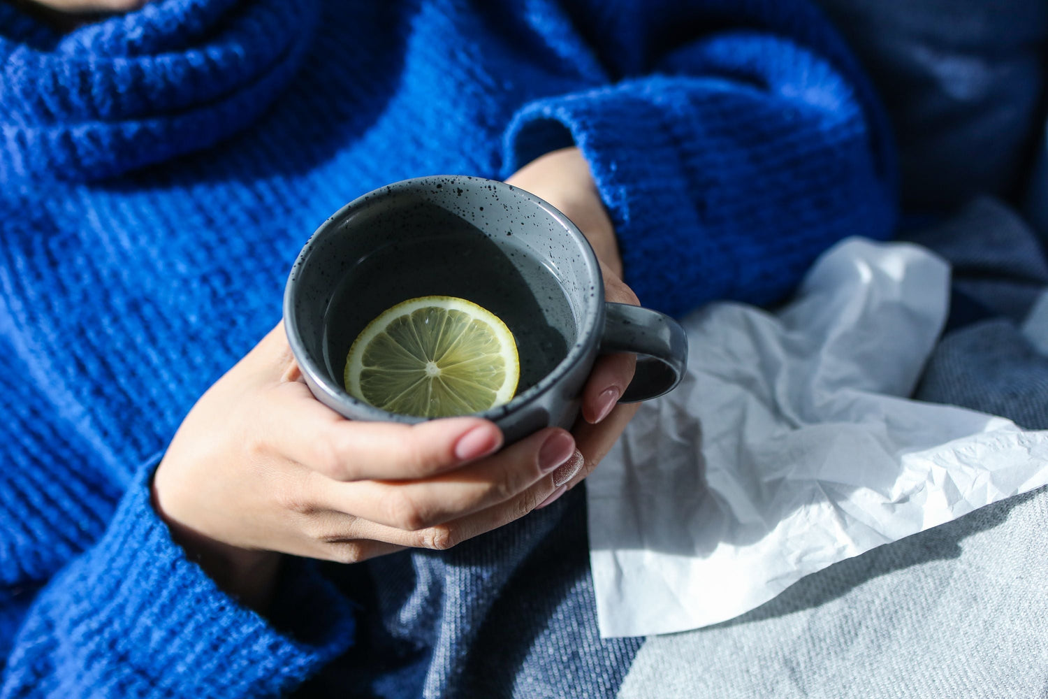 Person mit blauem Pullover hält graue Keramiktasse mit Zitronenscheibe, weiße Taschentücher im Hintergrund