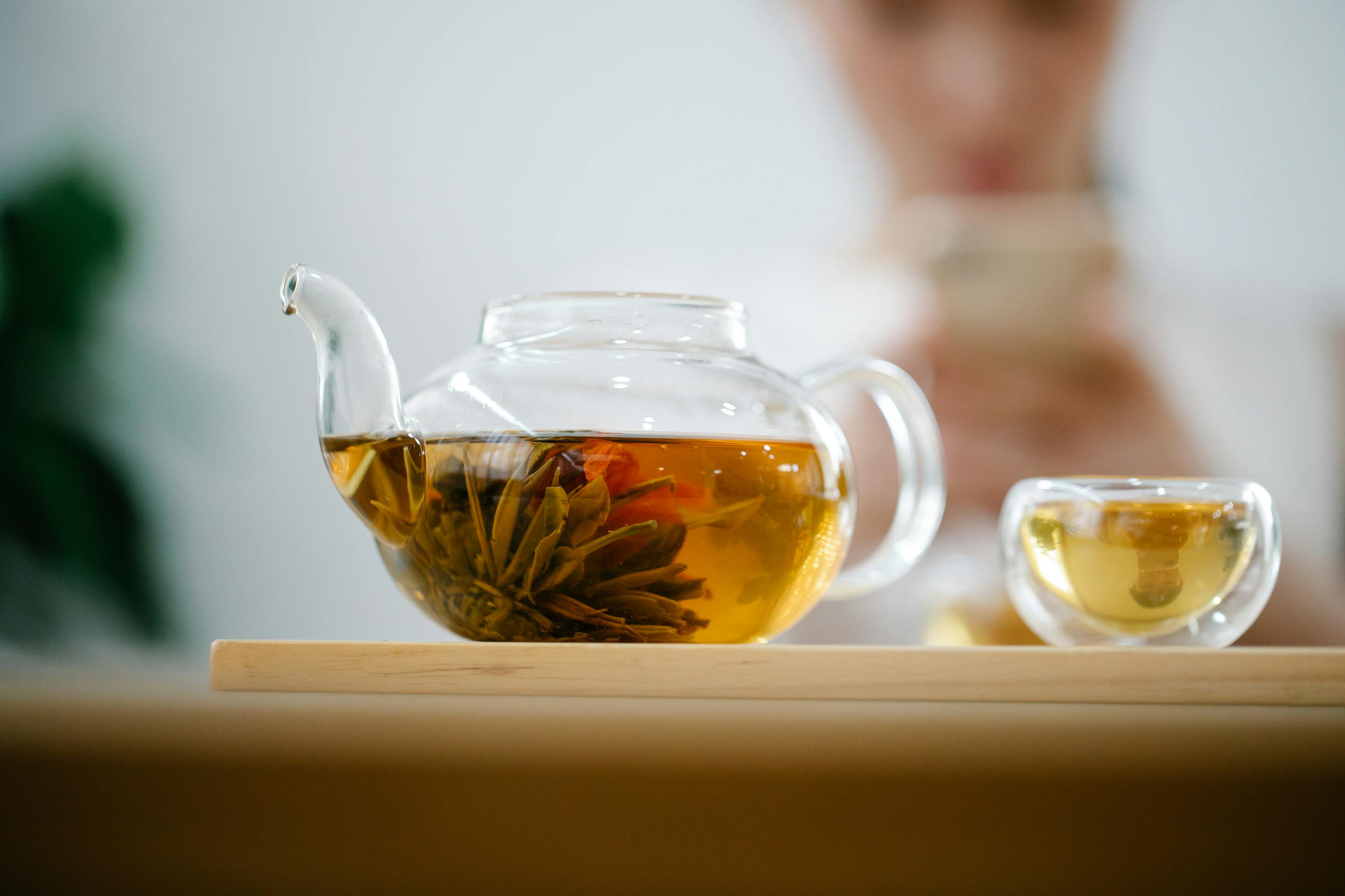 Glas-Teekanne mit blühender Teeblume und frisch aufgebrühtem Tee auf Holztablett