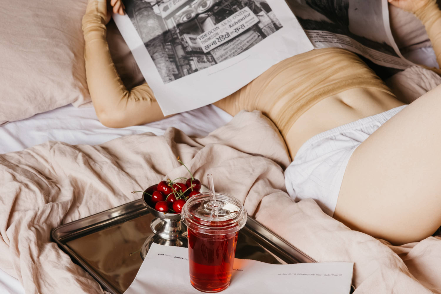Person entspannt auf einem Bett mit beigen Laken, liest eine Zeitung neben einem Tablett mit Kirschen und Getränk